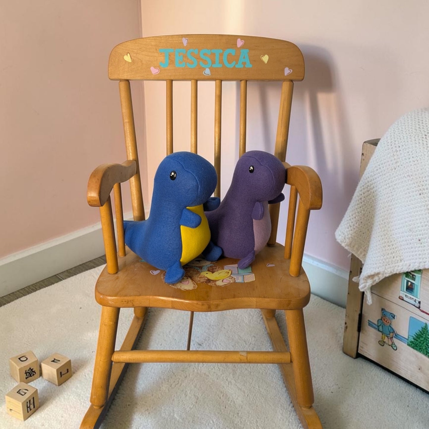 A wooden rocking chair in a nursery setting, featuring the name 'JESSICA' painted in blue on the backrest with small heart accents. Two plush dinosaur toys, one blue with a yellow belly and one purple, are sitting on the chair.