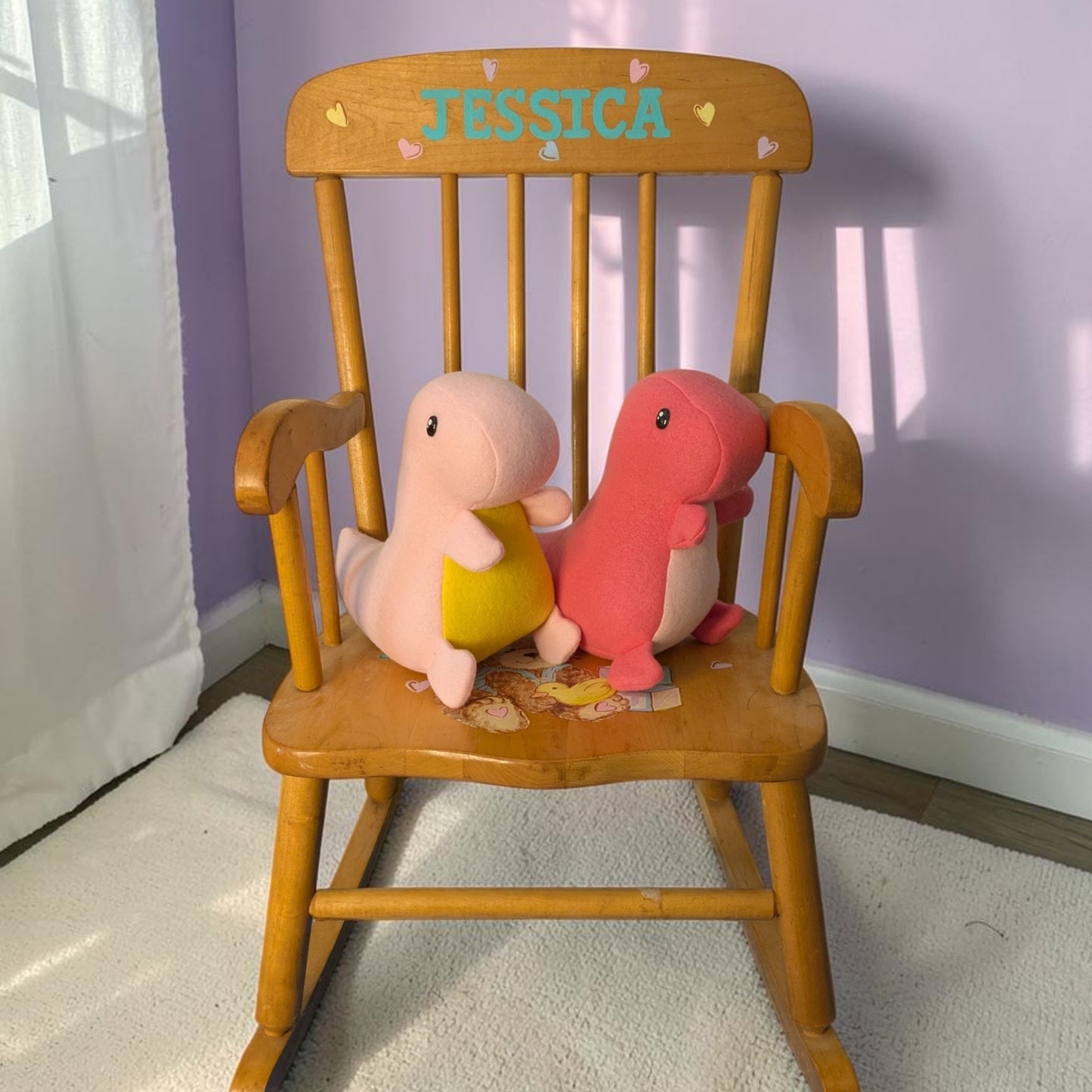 A wooden rocking chair with the name 'Jessica' and small hearts on the backrest, holding two plush dinosaur toys, one light pink with a yellow belly and one coral pink, in a room with a lavender wall and white curtain.