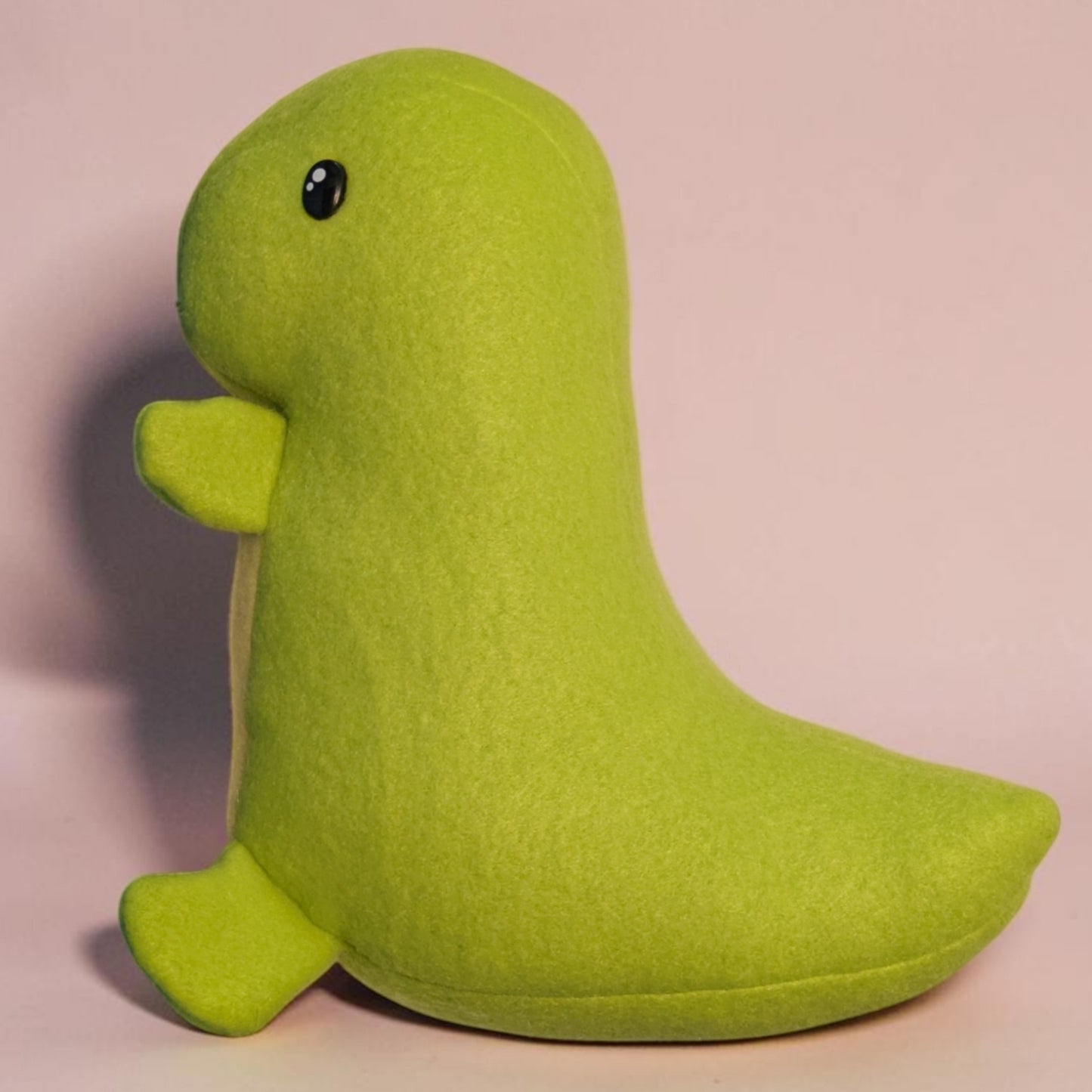 A lime green fleece plush toy shaped like a dinosaur, featuring a small black safety eye and tiny arms and legs, set against a solid light pink background.