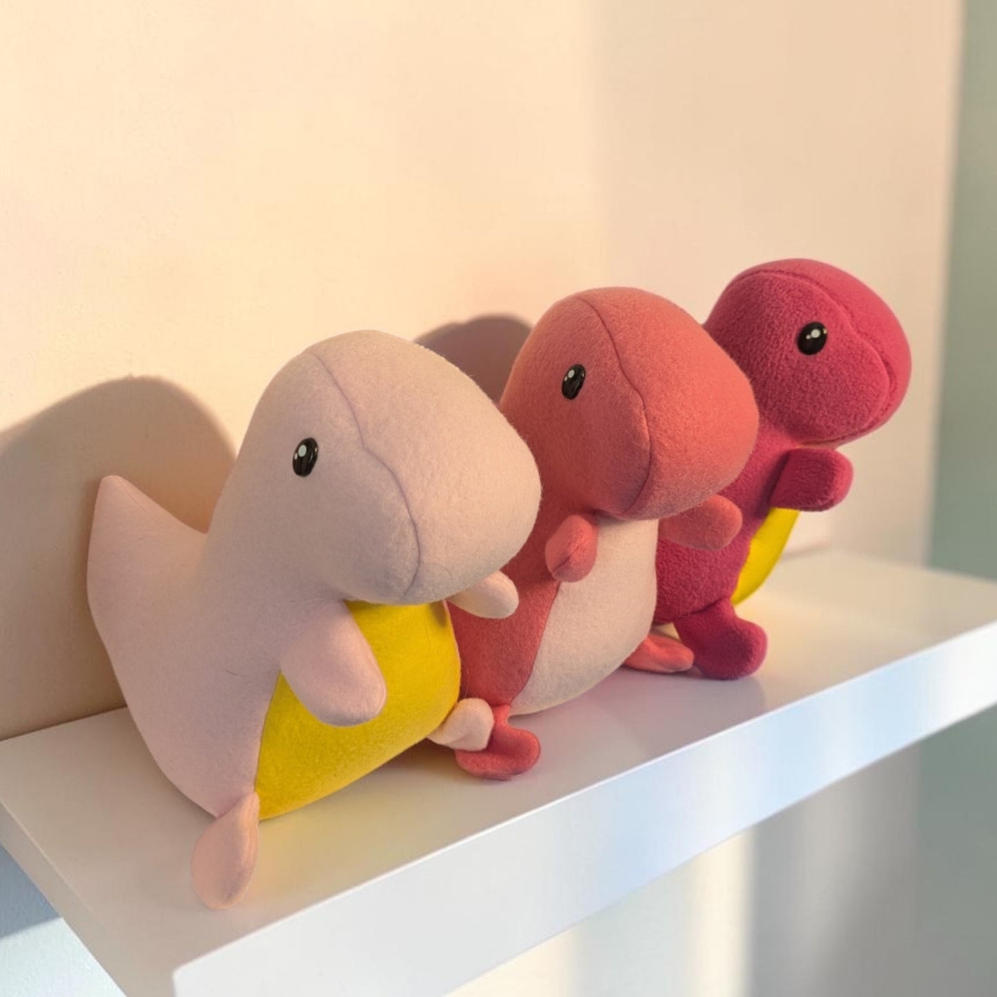 Three small plush dinosaur toys in pastel pink, coral pink, and magenta, each has a yellow belly, arranged on a white shelf against a softly lit beige wall.