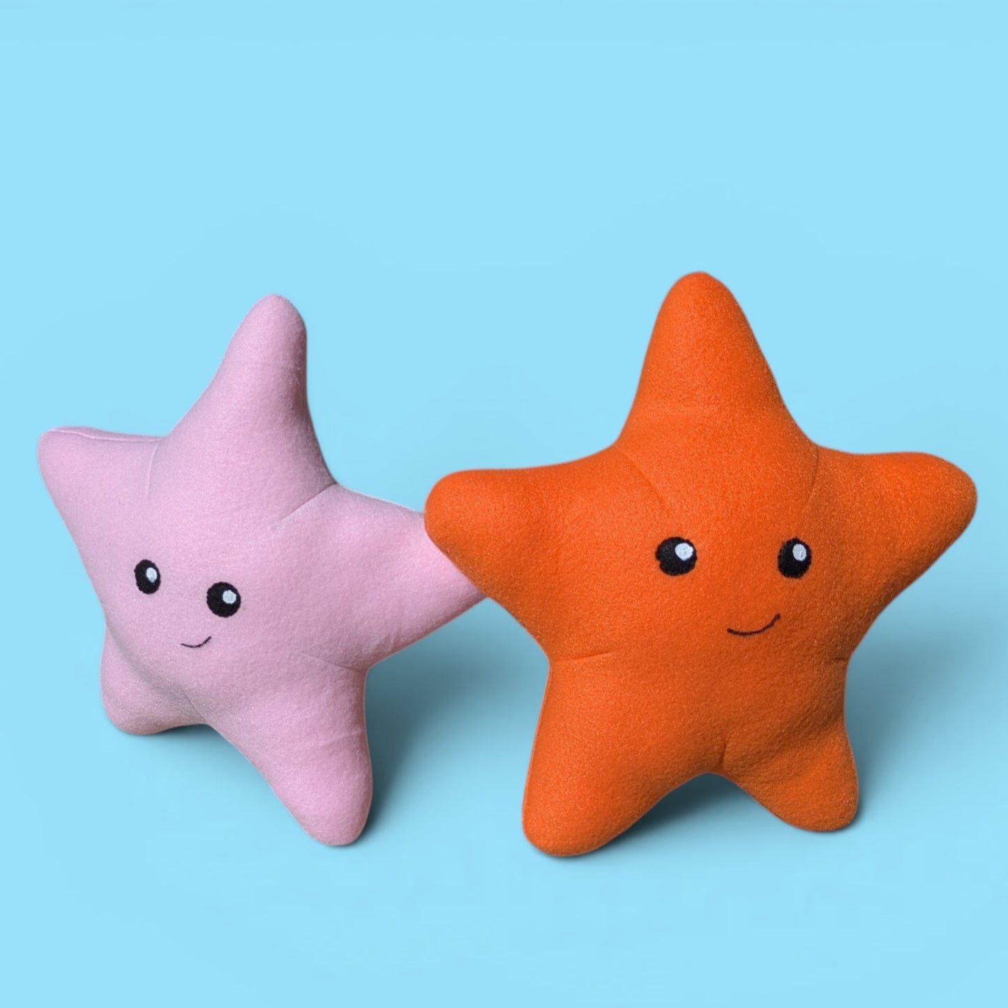 Two plush star-shaped stuffed animals on a light blue background, one in pink and one in orange, both with smiling faces.