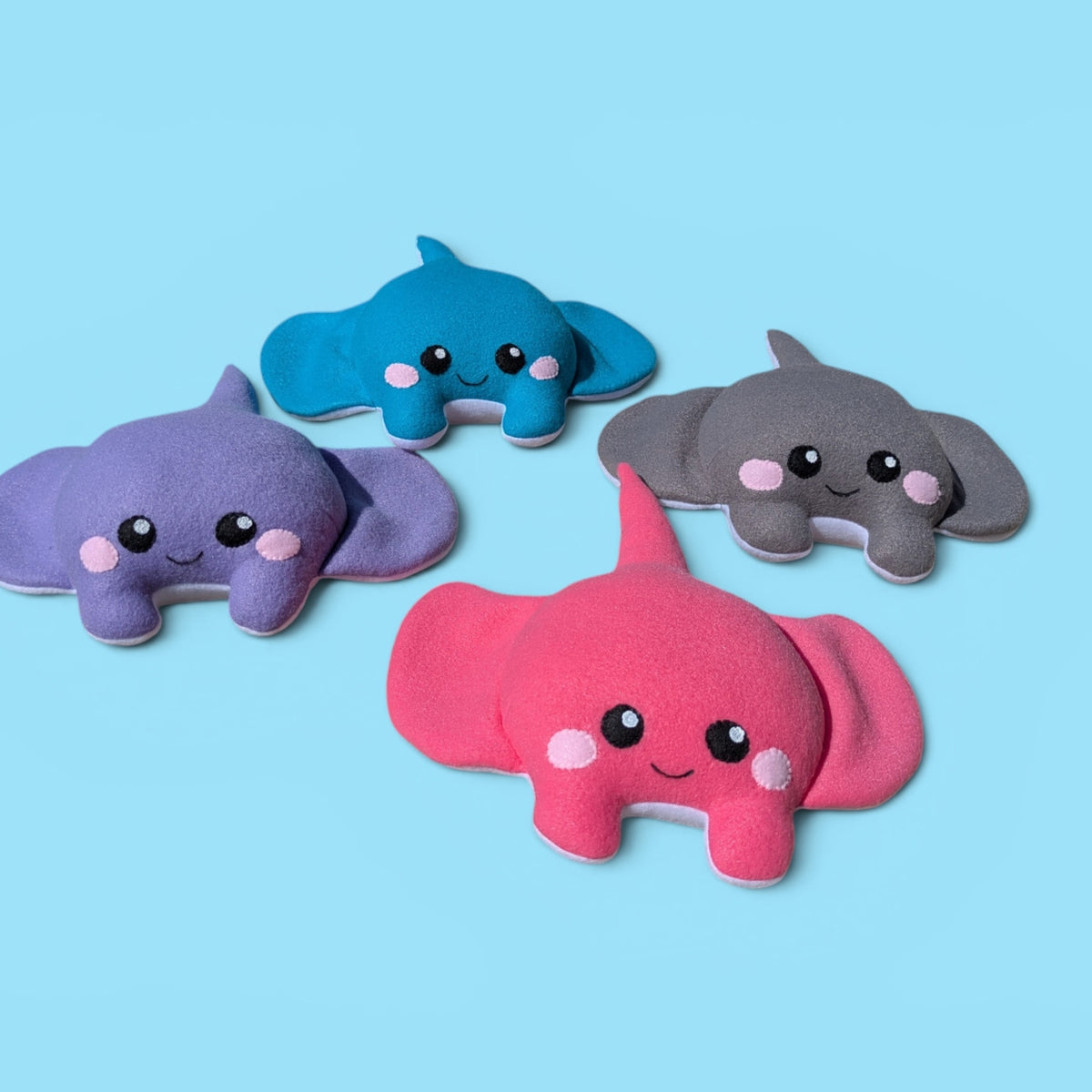 Four cute plush stingray toys in purple, blue, gray, and pink, arranged on a light blue background.