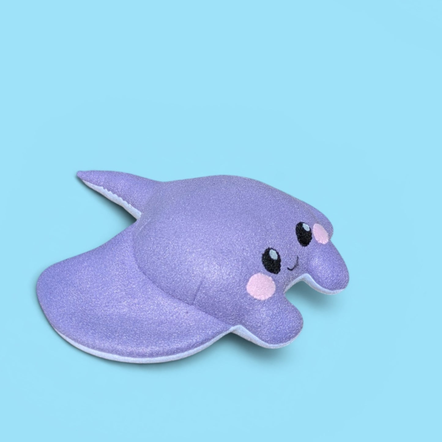 A cute, soft plush stingray toy in a soft lavender purple color, featuring felt black eyes, pink cheeks, and a small smile, set against a solid light blue background.