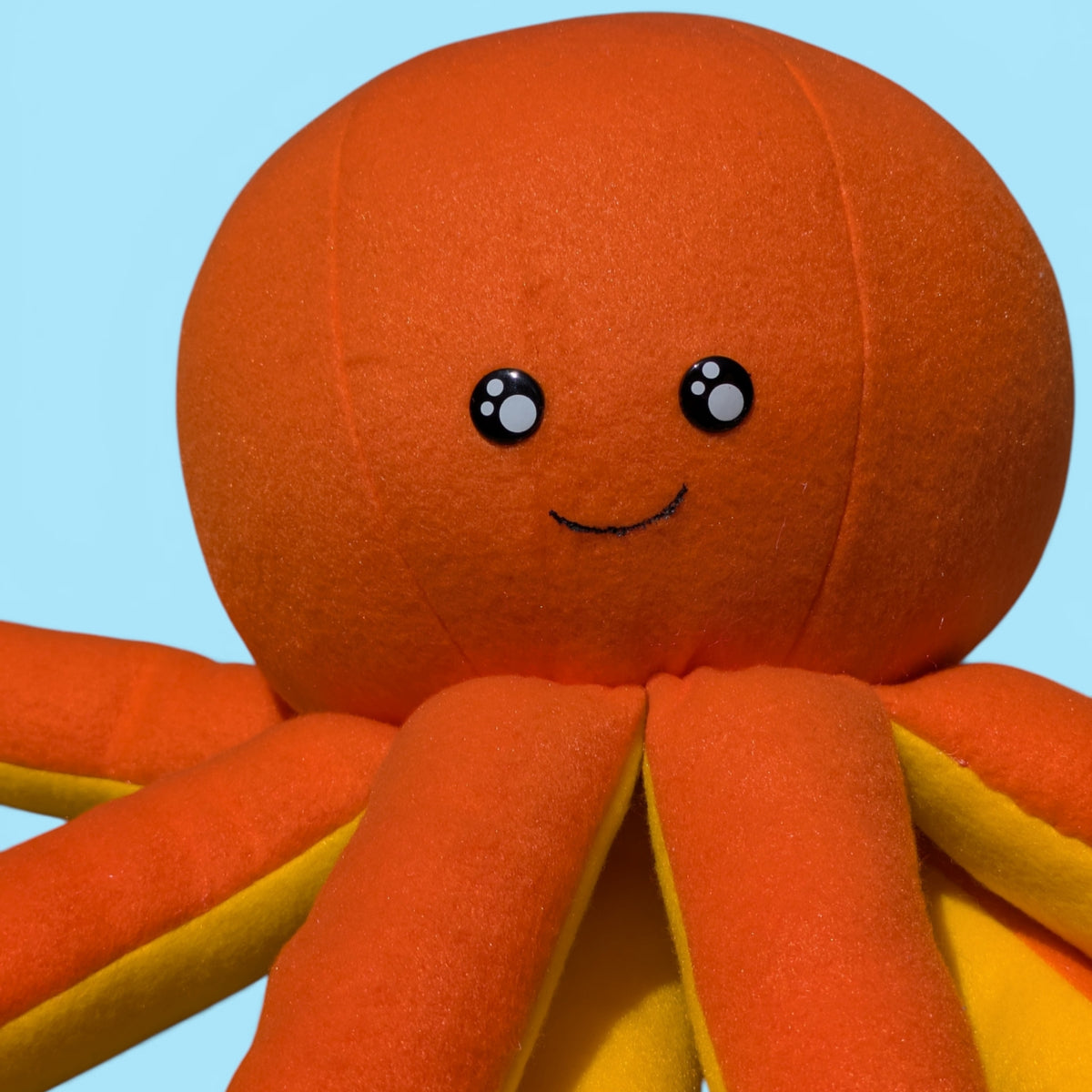 A close-up of a plush octopus toy with a round orange body, eight orange tentacles with yellow inner edges, and a simple face on a light blue background.