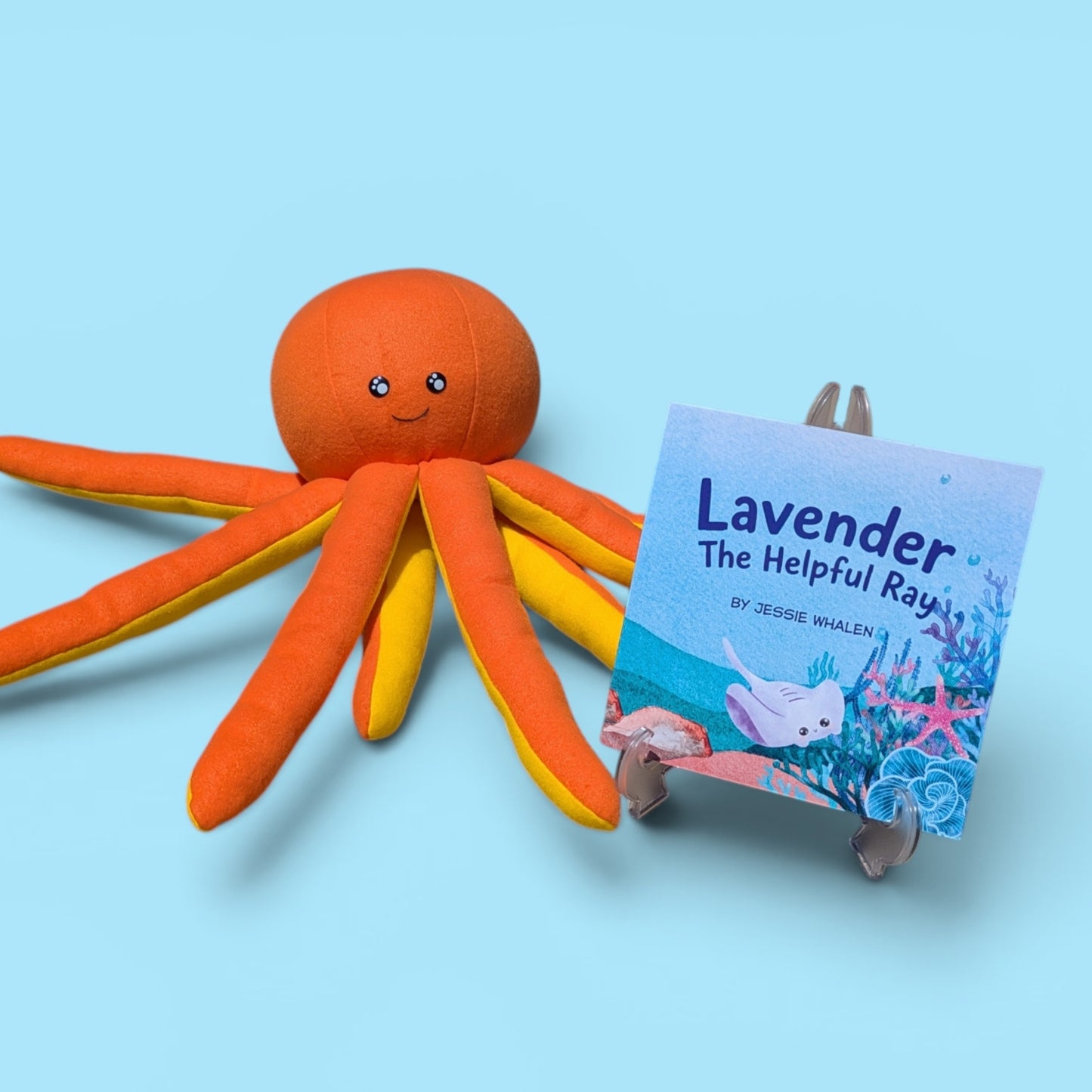 An orange octopus plush toy with yellow and orange striped tentacles sits beside a small book on a stand titled "Lavender The Helpful Ray" by Jessie Whalen, against a light blue background.