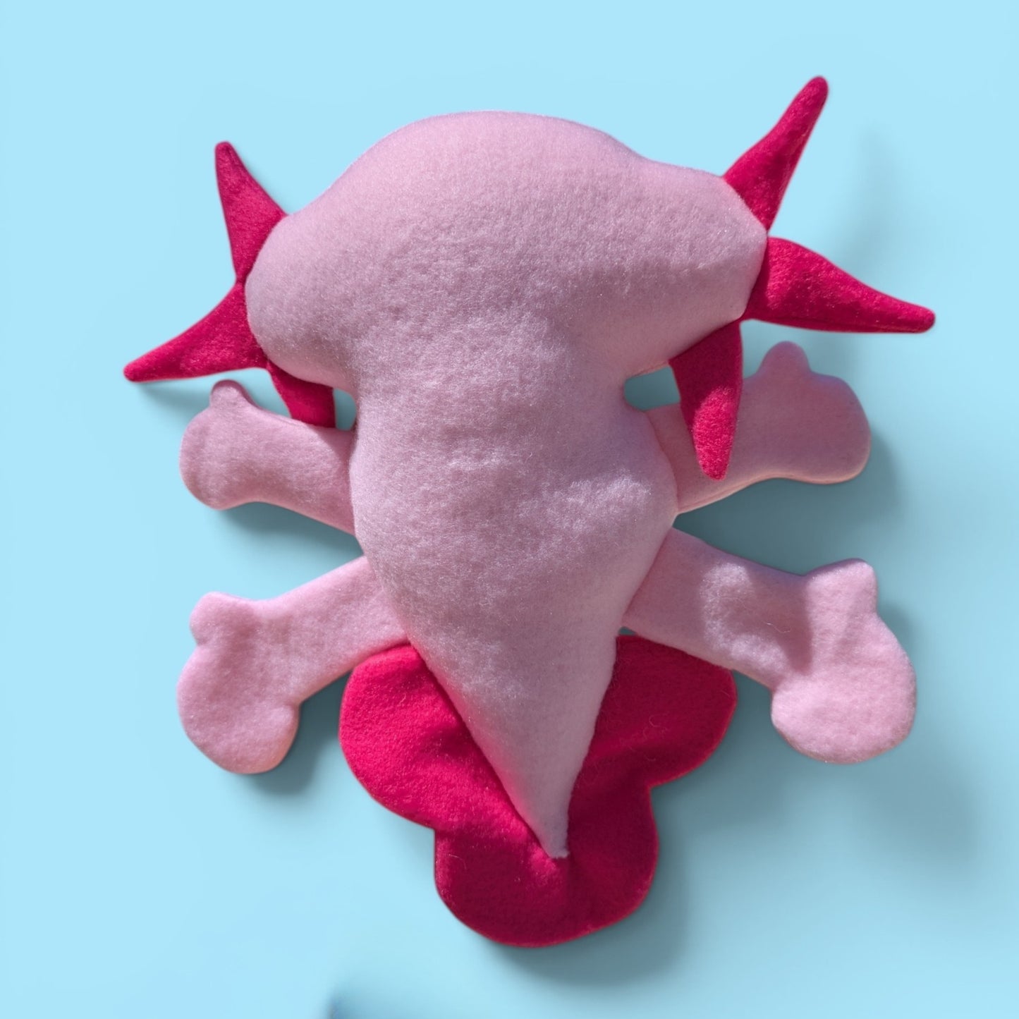 A pink and magenta plush toy shaped like a cartoon axolotl, photographed from the back on a light blue background.