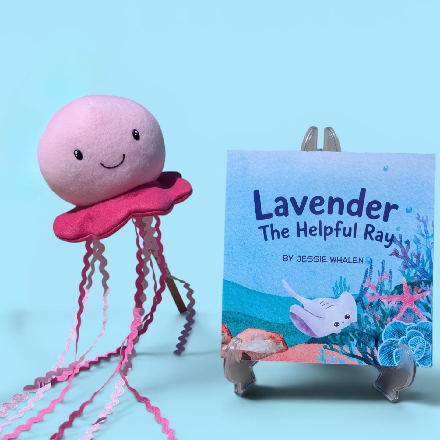 A pink plush jellyfish toy with long wavy pink tentacles stands beside a children's book titled "Lavender The Helpful Ray" by Jessie Whalen, displayed on a small stand against a light blue background.