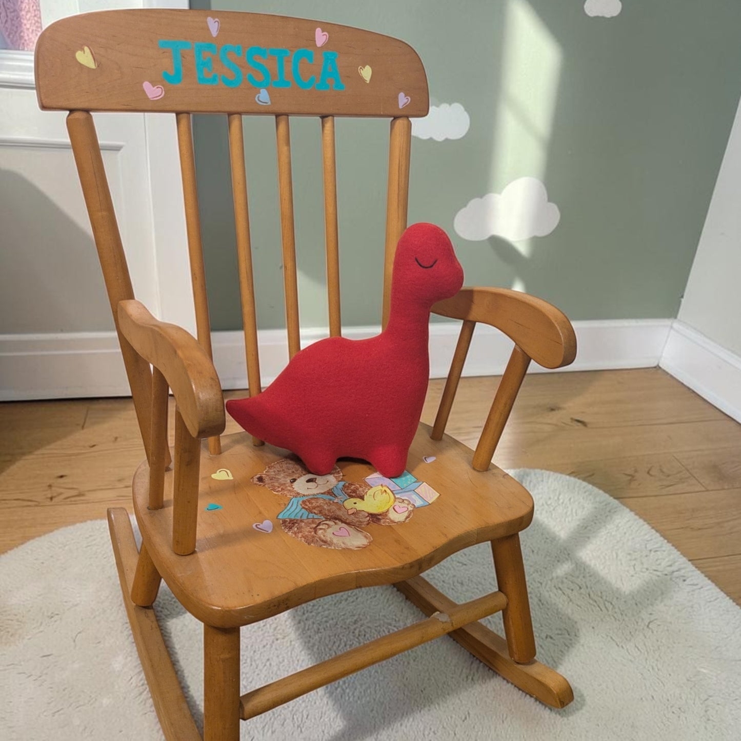 A wooden children's rocking chair with 'JESSICA' personalized on the backrest, featuring a red plush dinosaur toy on the seat, set in a child's room.