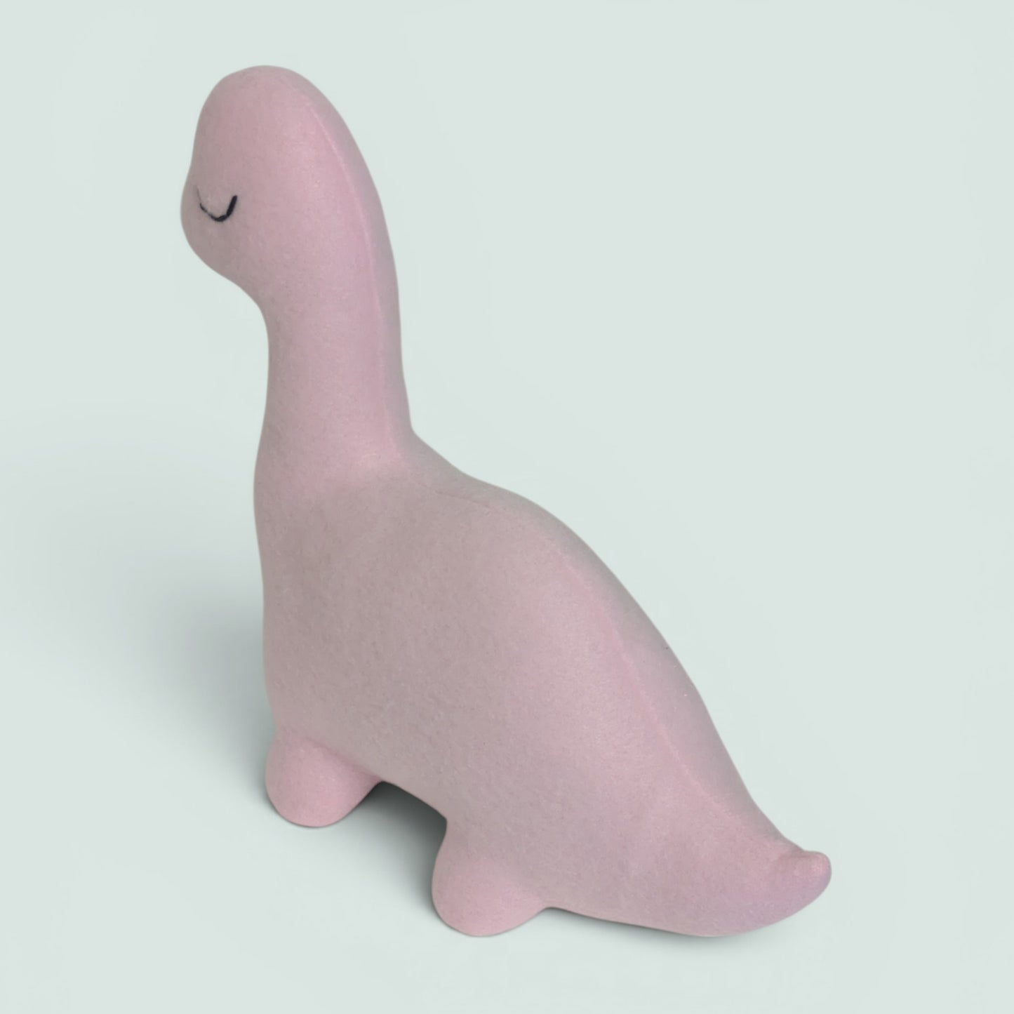 A soft, matte pink dinosaur toy with a long neck and closed eyes, shown on a clean, light gray background.