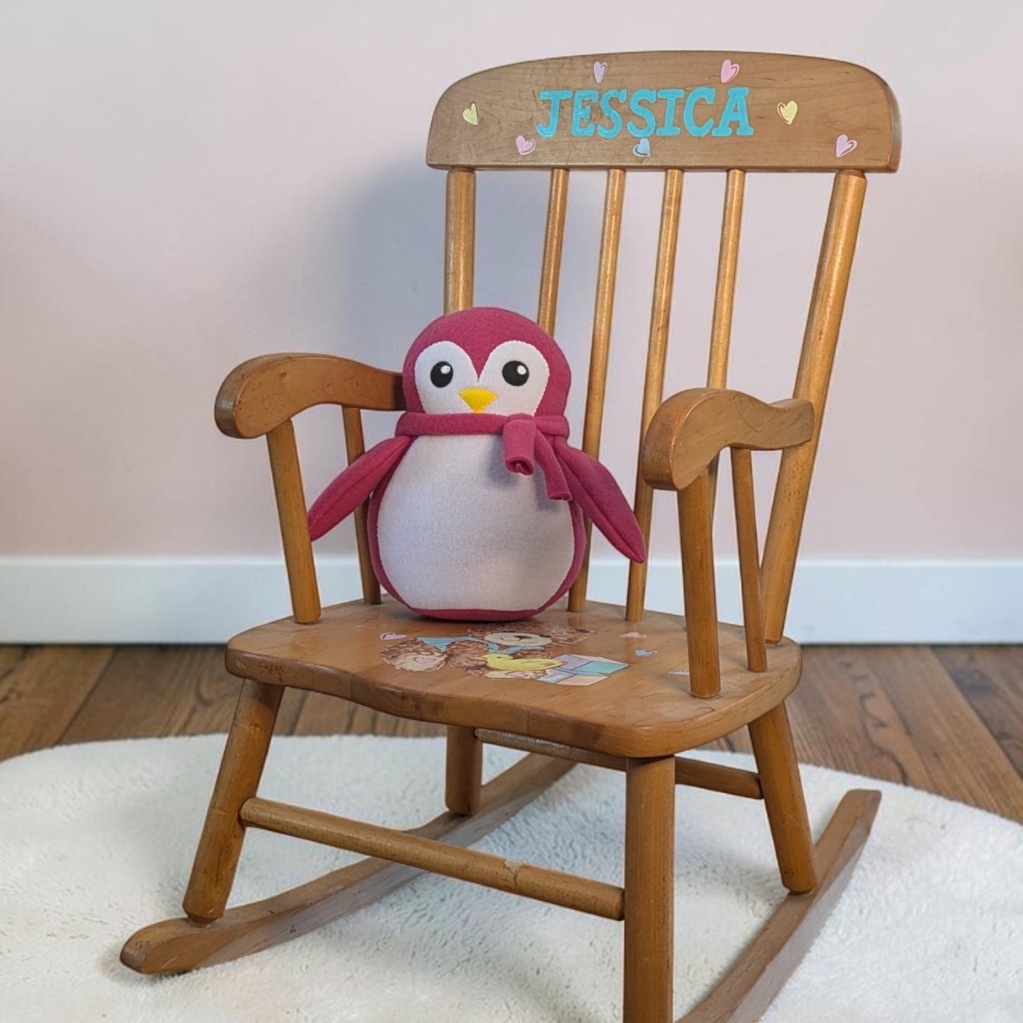 A wooden rocking chair with a custom name 'JESSICA' on the backrest, featuring a plush penguin toy sitting on the seat. The chair is placed on a white rug over a wooden floor against a light pink wall.