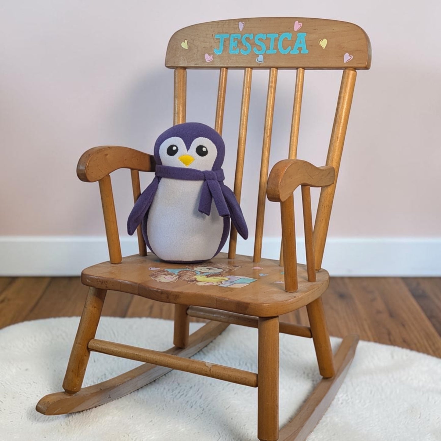 A wooden rocking chair with the name 'JESSICA' and small hearts painted on the backrest, holding a plush penguin toy with a purple scarf. The chair is placed on a white rug over a wooden floor against a light wall.
