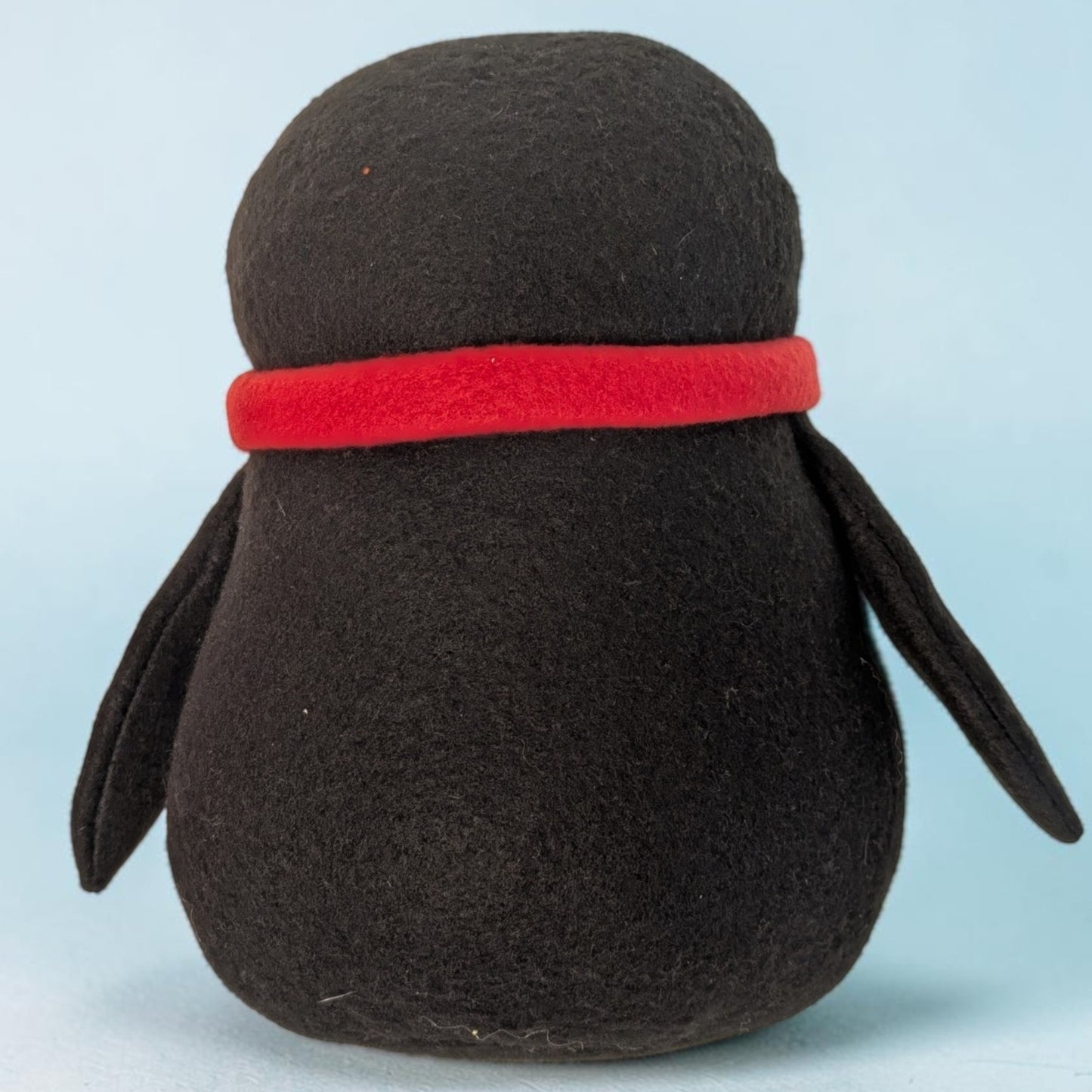 A black fleece penguin plush toy with a bright red felt scarf, shown from the back against a light blue background.