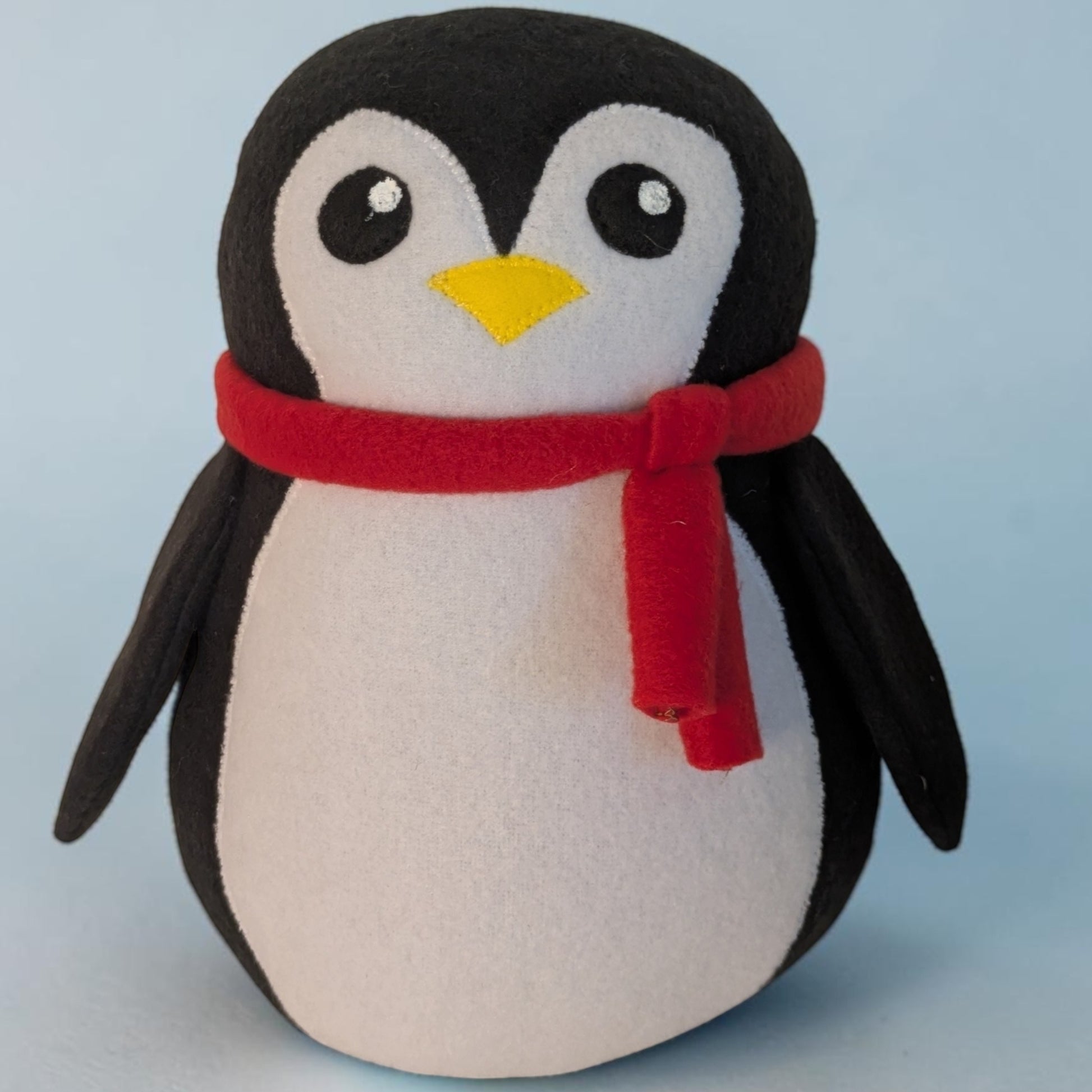 A plush penguin toy with black and white feathers, a small yellow beak, and a red scarf, photographed against a light blue background.