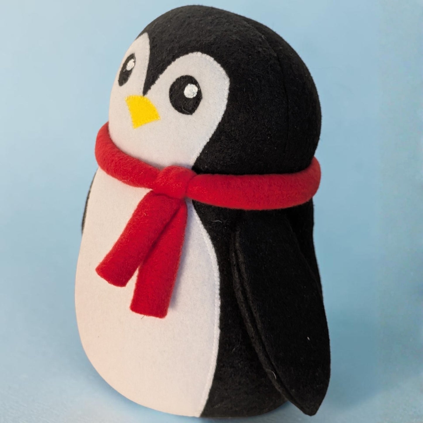 A plush penguin toy with black and white fleece body, a yellow beak, and a red scarf, photographed against a light blue background.