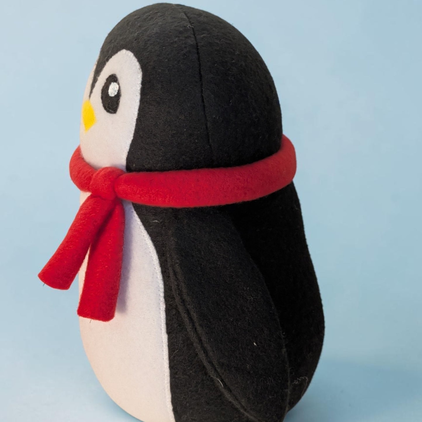 A plush penguin toy made from fleece fabric, featuring a black back, white belly, a small yellow beak, and a red scarf, set against a solid light blue background.