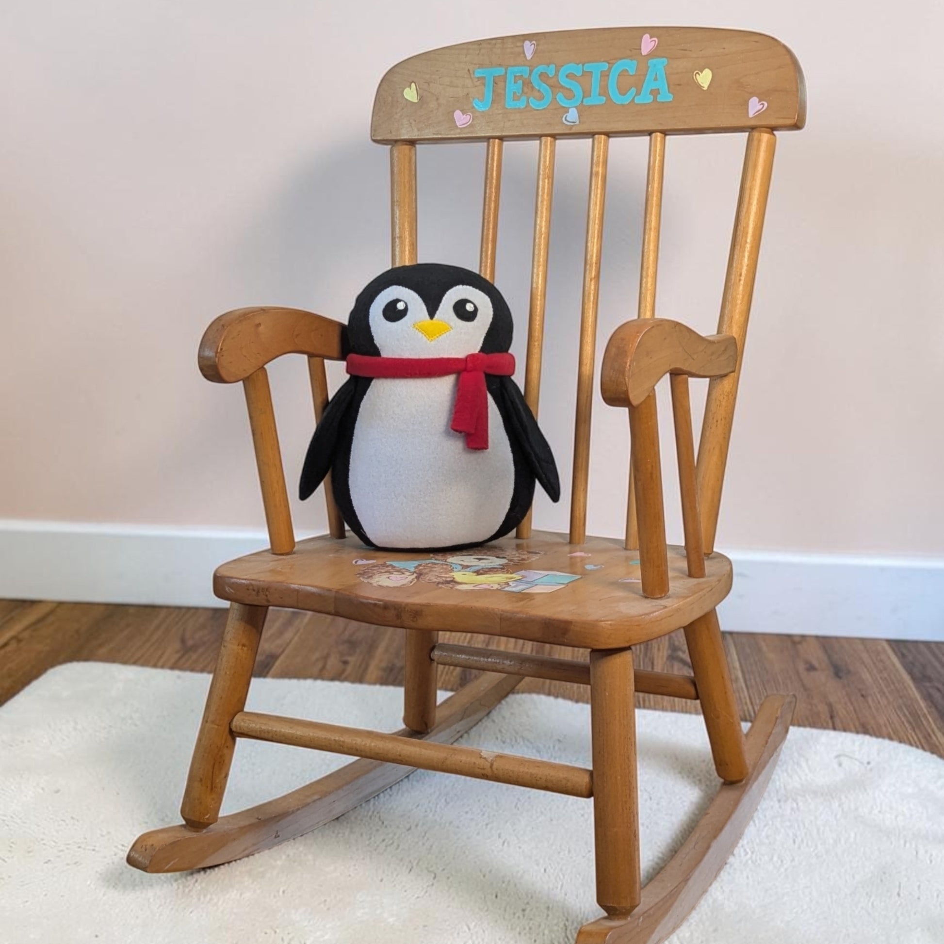 A wooden rocking chair with the name 'JESSICA' painted on the backrest, styled with a plush penguin toy wearing a red scarf sitting on the seat, set on a white rug over a wooden floor.