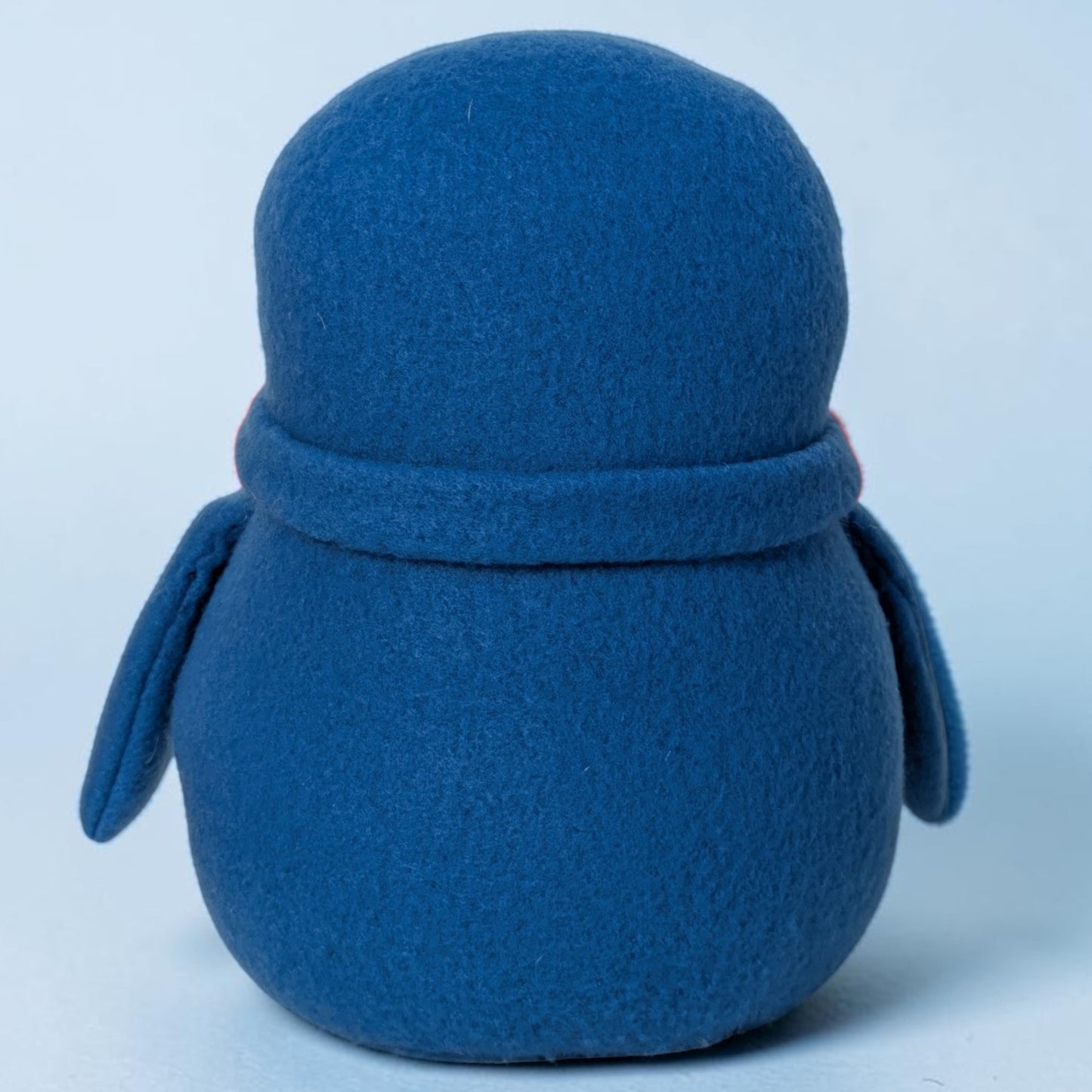 A blue fleece penguin stuffed animal photographed from the back against a light blue studio background.