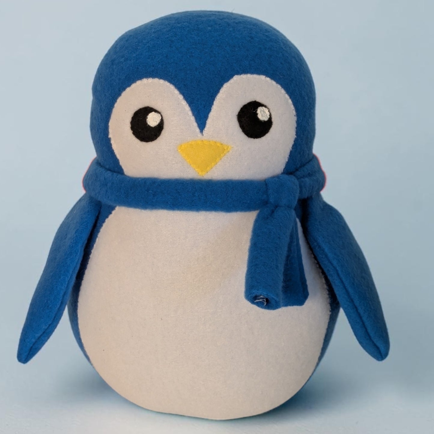 A plush penguin toy with a blue body, white face and belly, black eyes, a yellow beak, and a blue scarf, photographed against a light blue background.