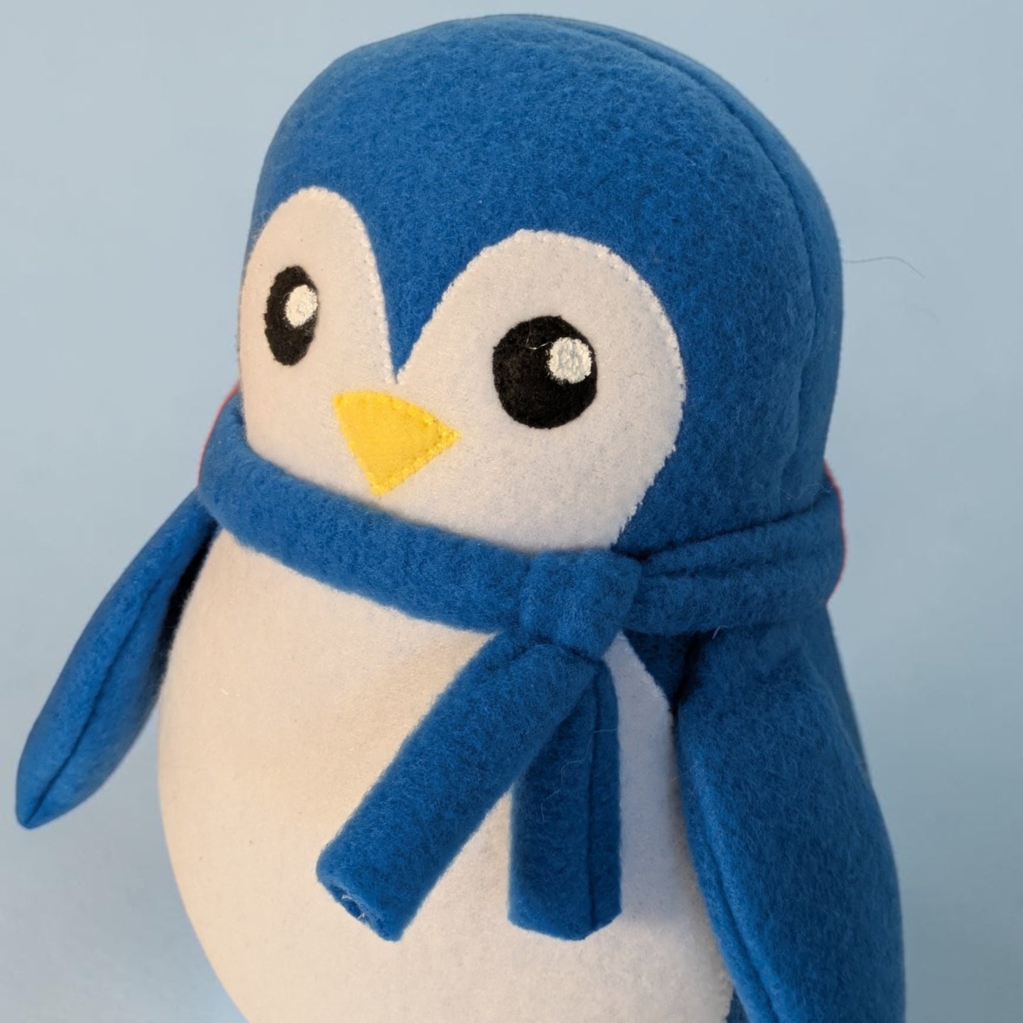 A blue and white plush penguin toy wearing a matching blue scarf, photographed against a light blue background.