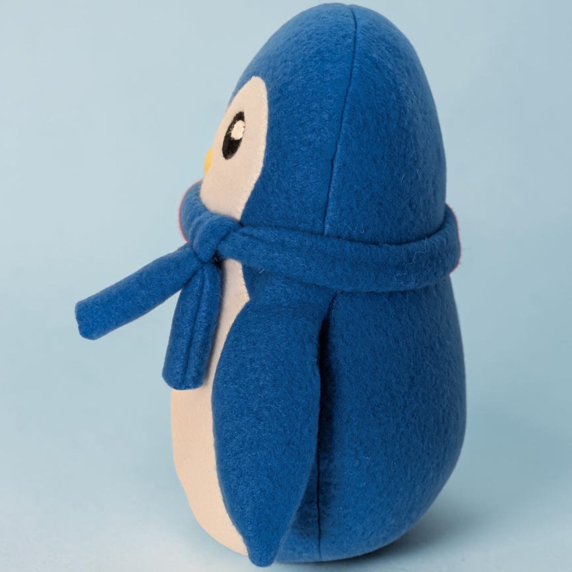 A blue plush penguin toy wearing a matching blue scarf, photographed against a light blue studio background.