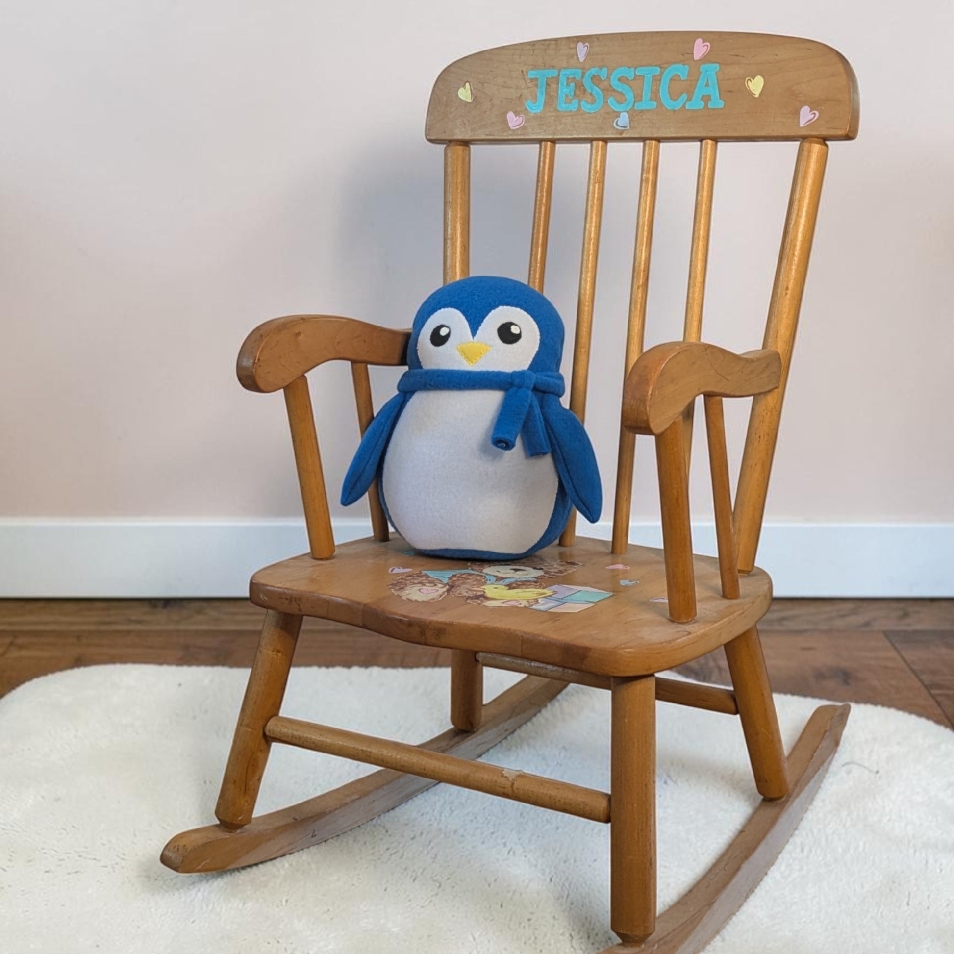 A wooden rocking chair with the name 'JESSICA' painted in blue on the backrest, decorated with small hearts and styled with a blue and white penguin plush toy sitting on the seat.