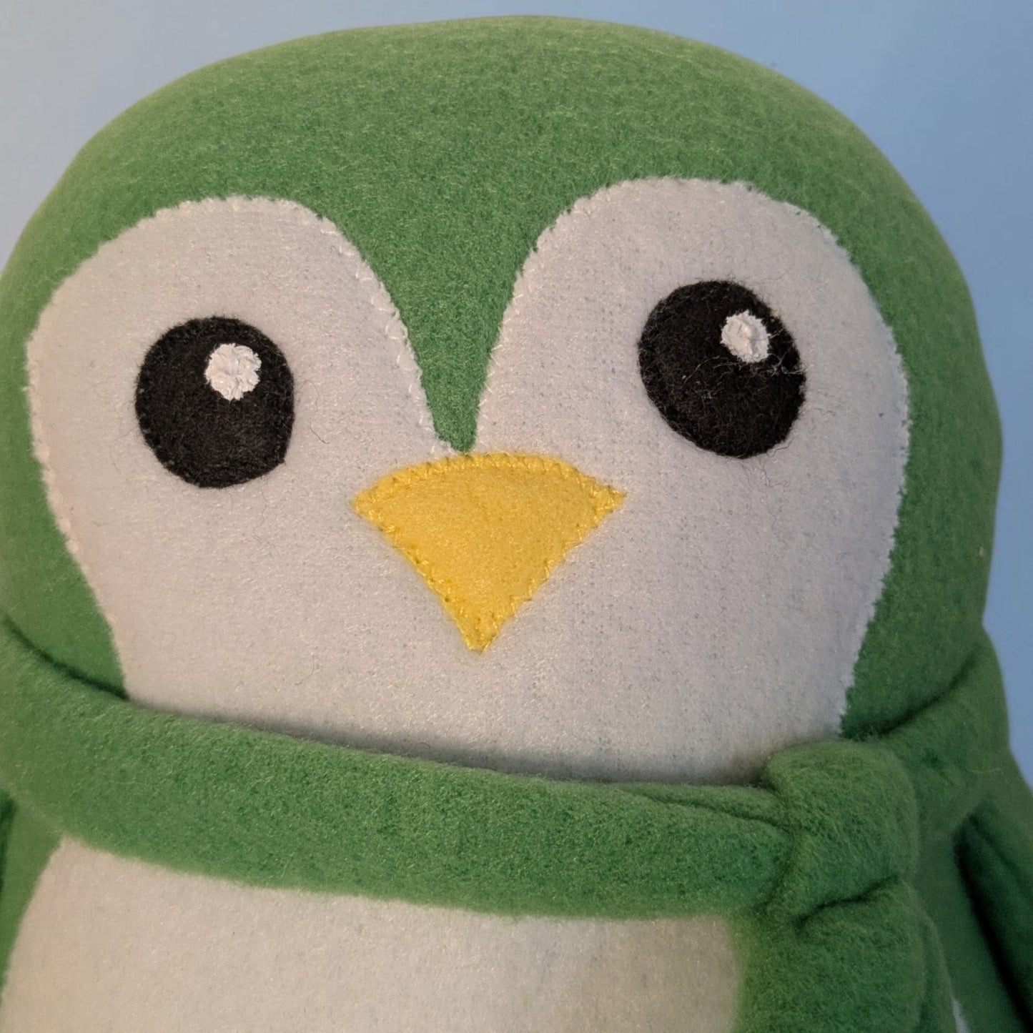 A close-up of a green and white plush penguin toy wearing a green scarf, with black felt eyes and a yellow beak, against a light blue background.