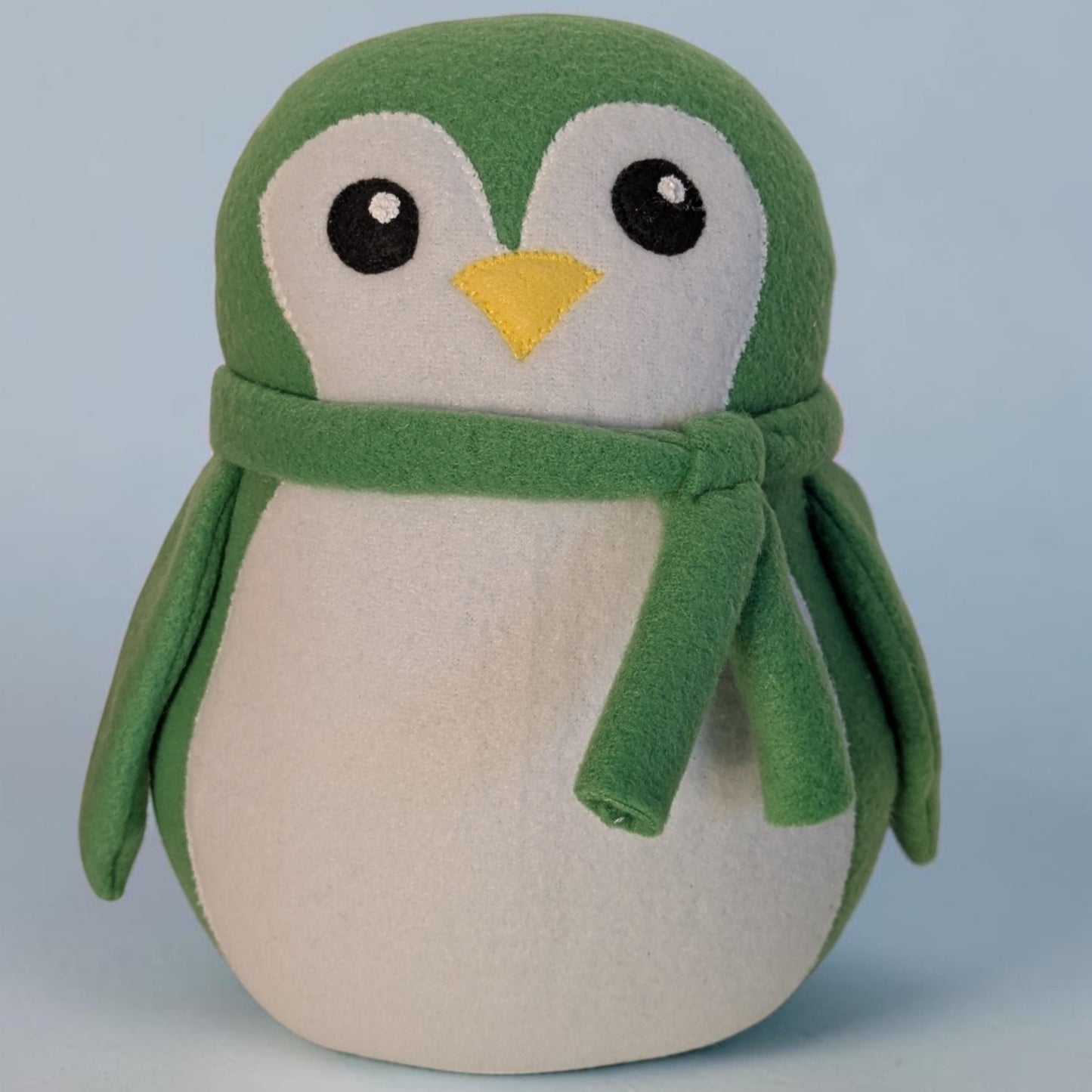 A plush penguin stuffed animal toy with a green fleece head, wings, and scarf, a cream-colored belly and face, and a small yellow beak, set against a light blue background.