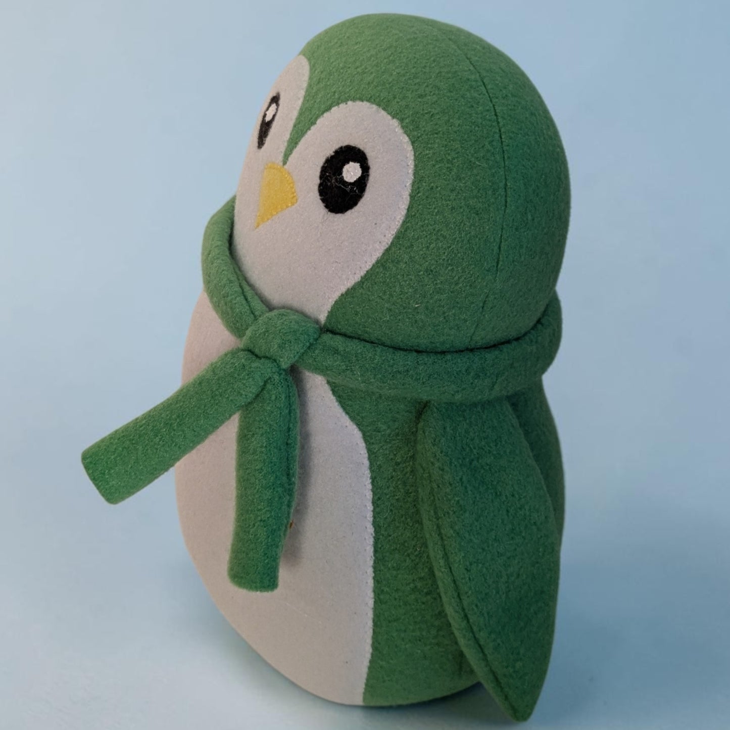 A green and white plush penguin toy wearing a matching green scarf, photographed against a soft light blue background.