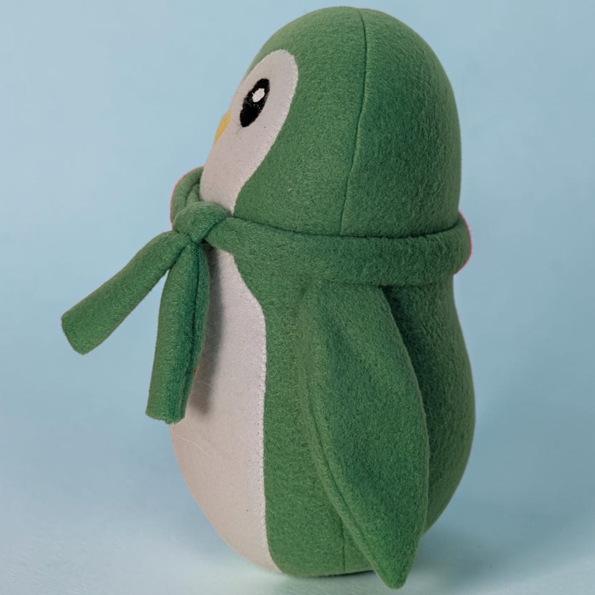 A green penguin plush toy wearing a matching green scarf, photographed against a light blue studio background.