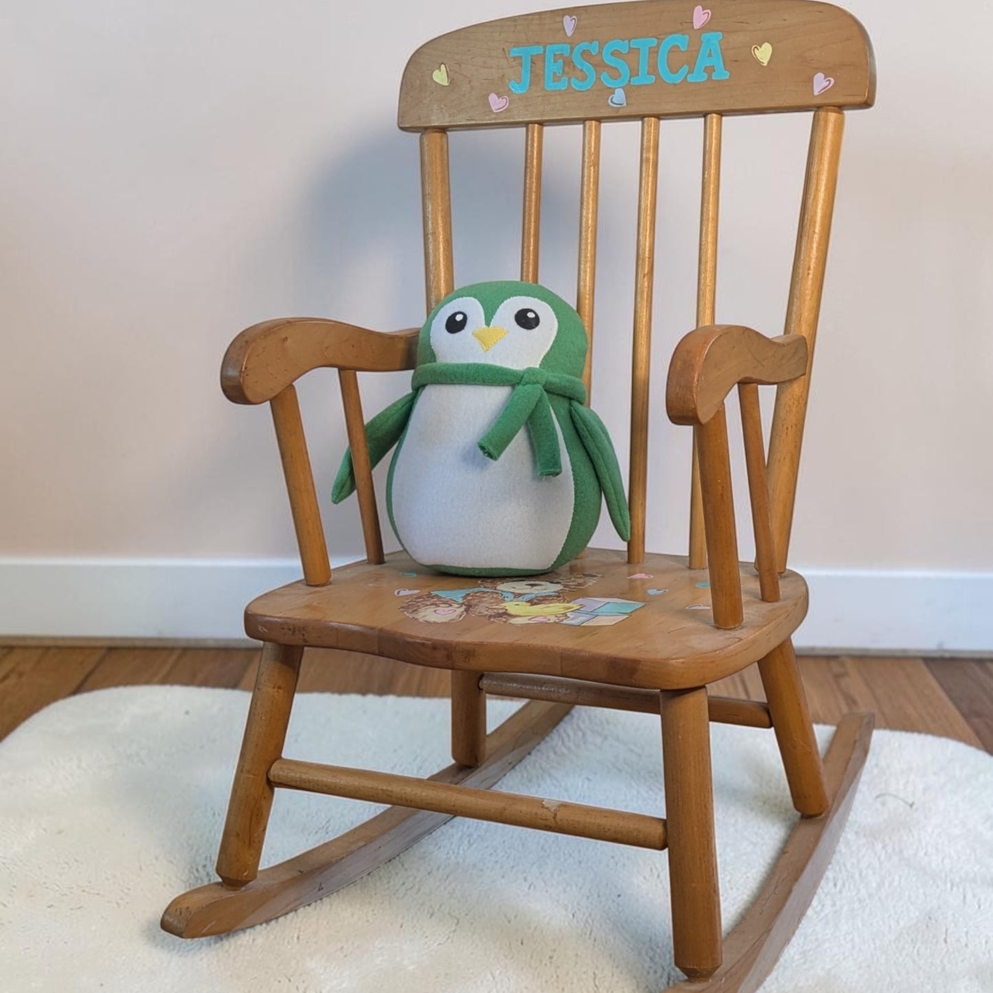 A wooden rocking chair with the name 'JESSICA' painted in blue on the backrest, featuring a green and white penguin plush toy sitting on the seat. The chair is placed on a white textured rug over a wooden floor against a plain light wall.