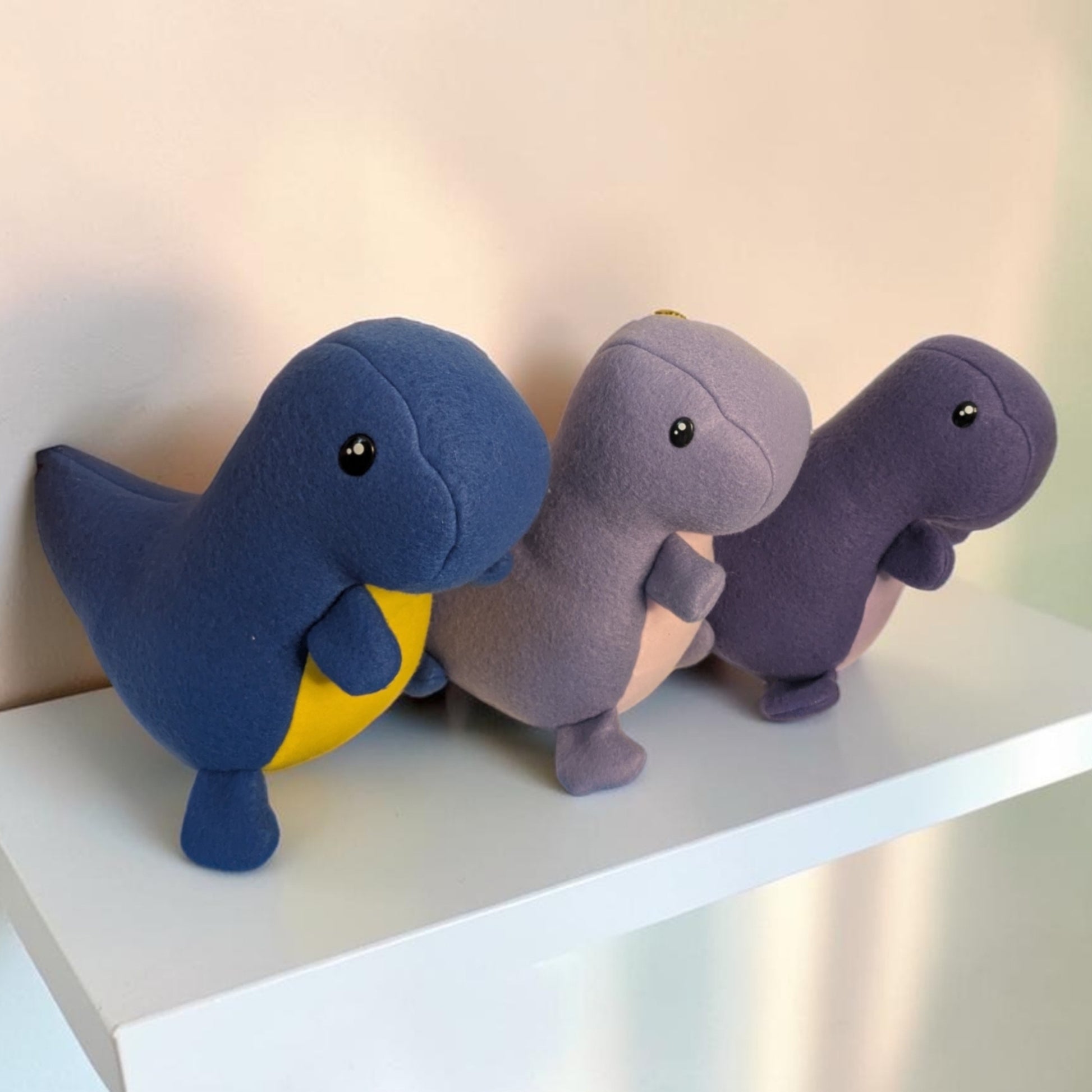 Three plush dinosaur toys lined up on a white shelf against a light beige wall. The left dinosaur is blue with a yellow belly, the middle one is lavender with a pale pink underside, and the right one is dark purple with a pale pink underside.