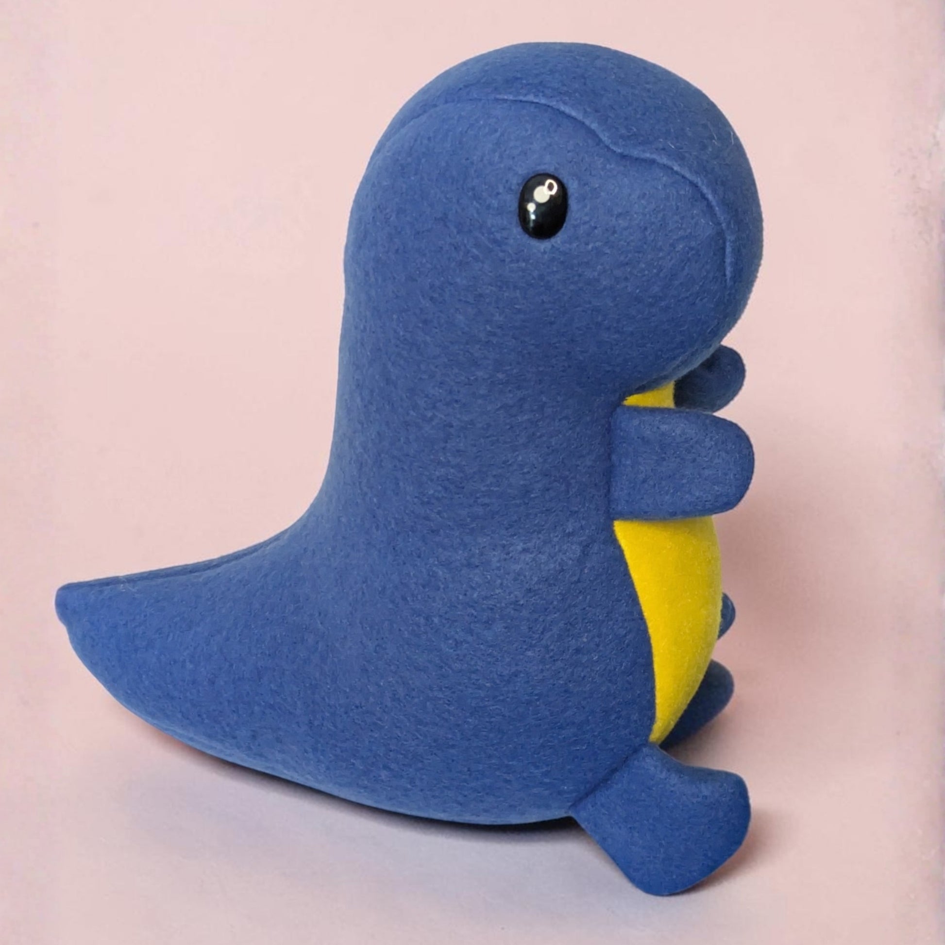 A blue fleece dinosaur plush toy with a yellow belly and black safety eyes, sitting against a soft pink background.