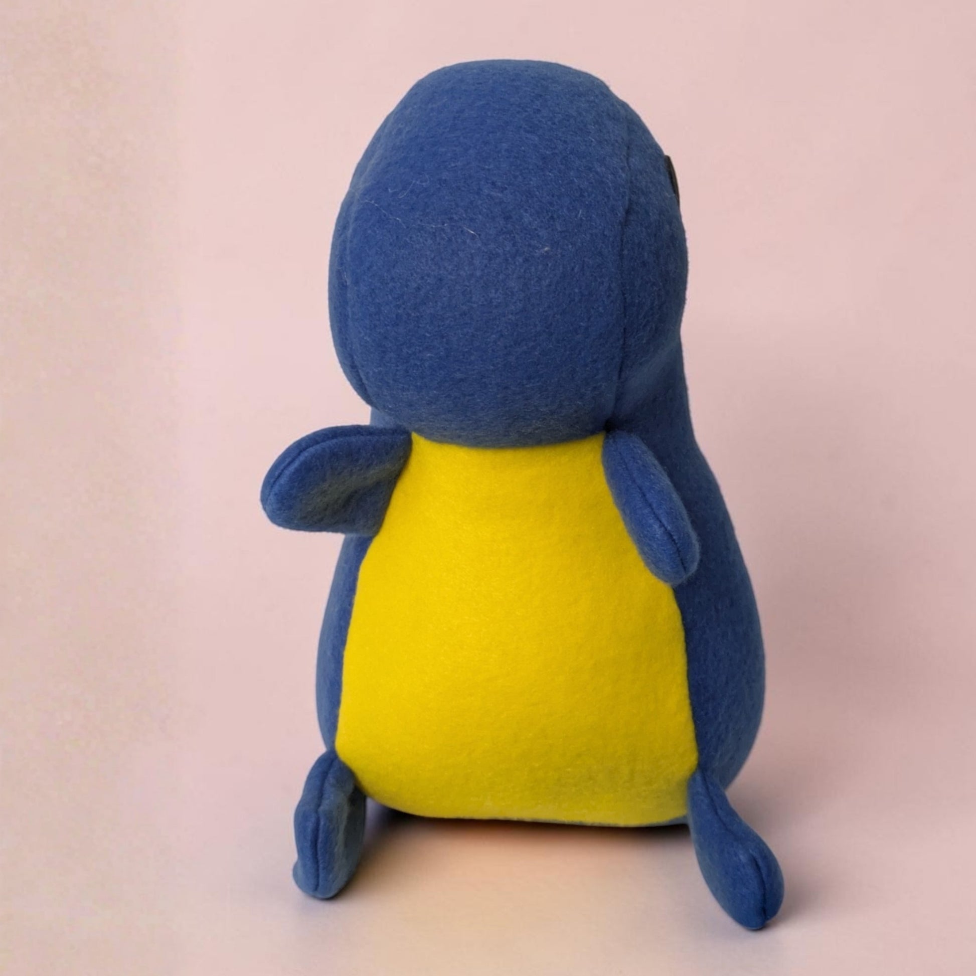 A blue and yellow plush toy shaped like a dinosaur, shown from the front against a soft pink background.