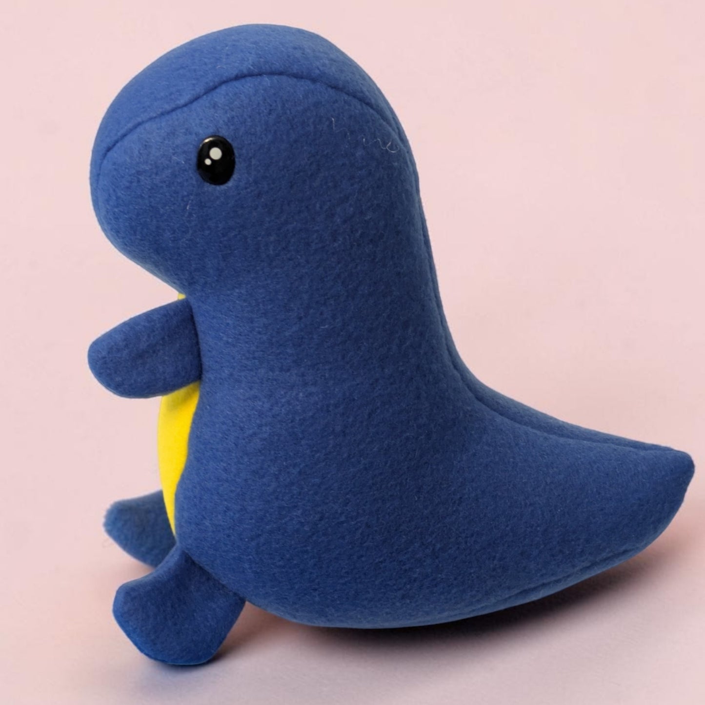 A blue plush dinosaur toy with a small yellow belly patch and black safety eyes, photographed against a solid pale pink background.