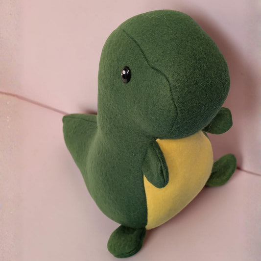 A green plush dinosaur toy with a yellow belly and small black safety eyes, sitting against a soft pink background.