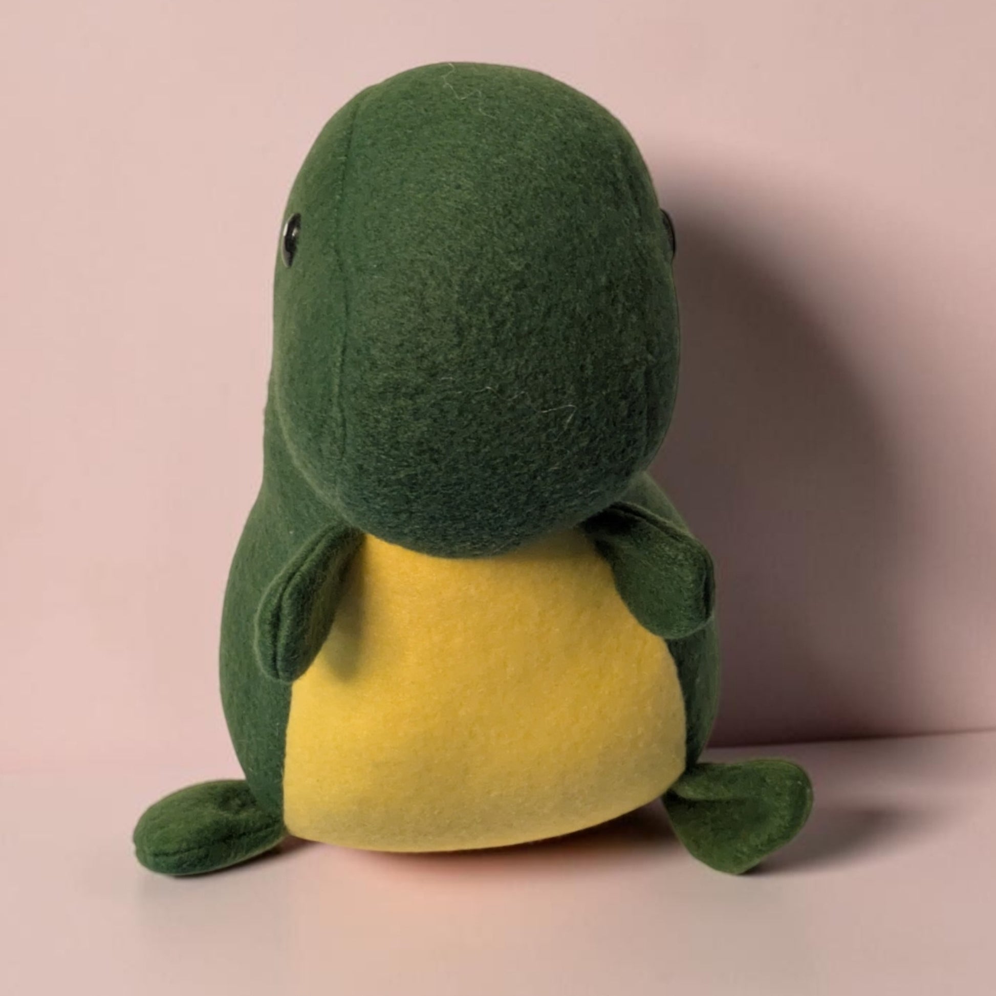 A green and yellow plush dinosaur toy with a soft fleece texture, sitting against a pale pink background.