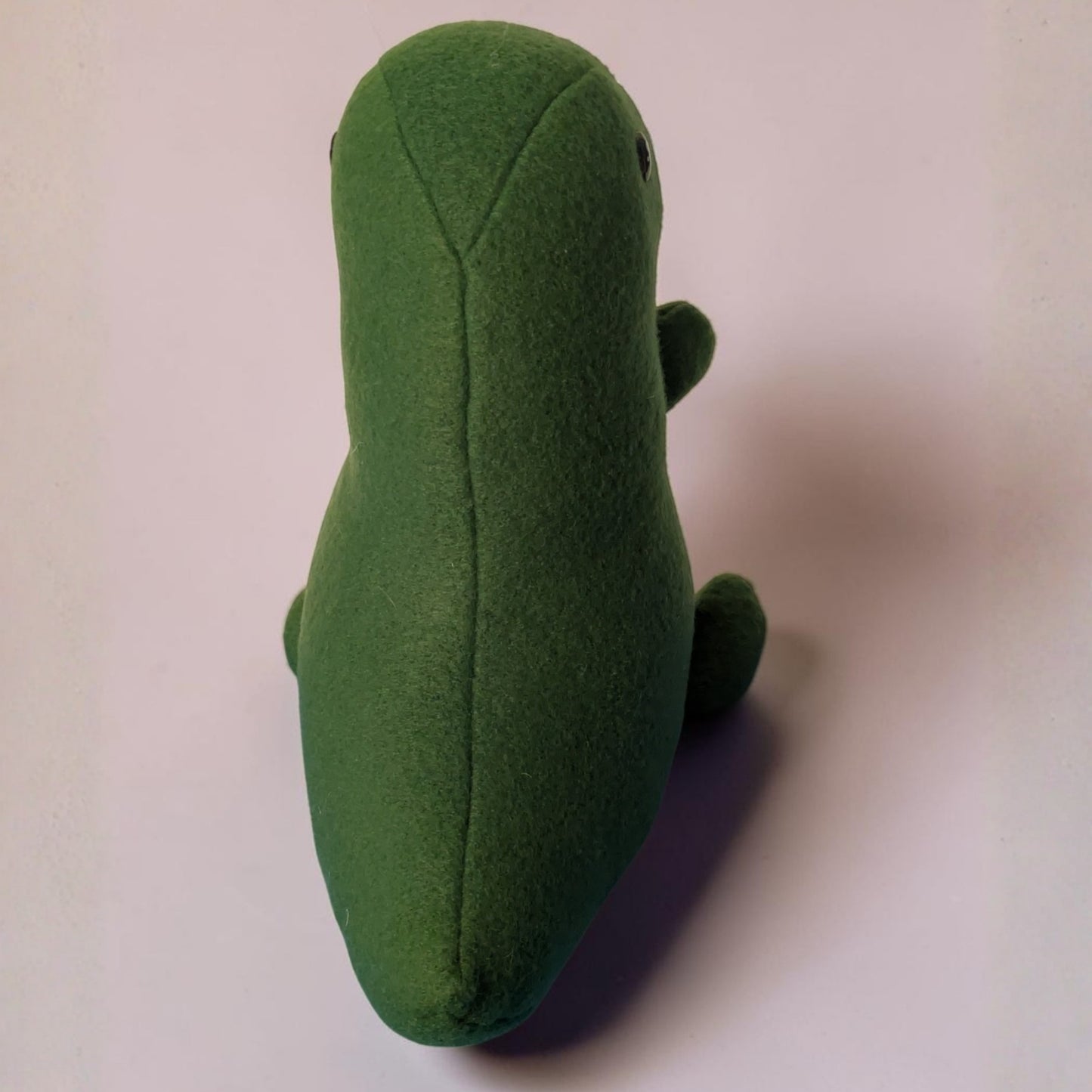 A green plush toy shaped like a stylized dinosaur, photographed from the back on a plain light background.