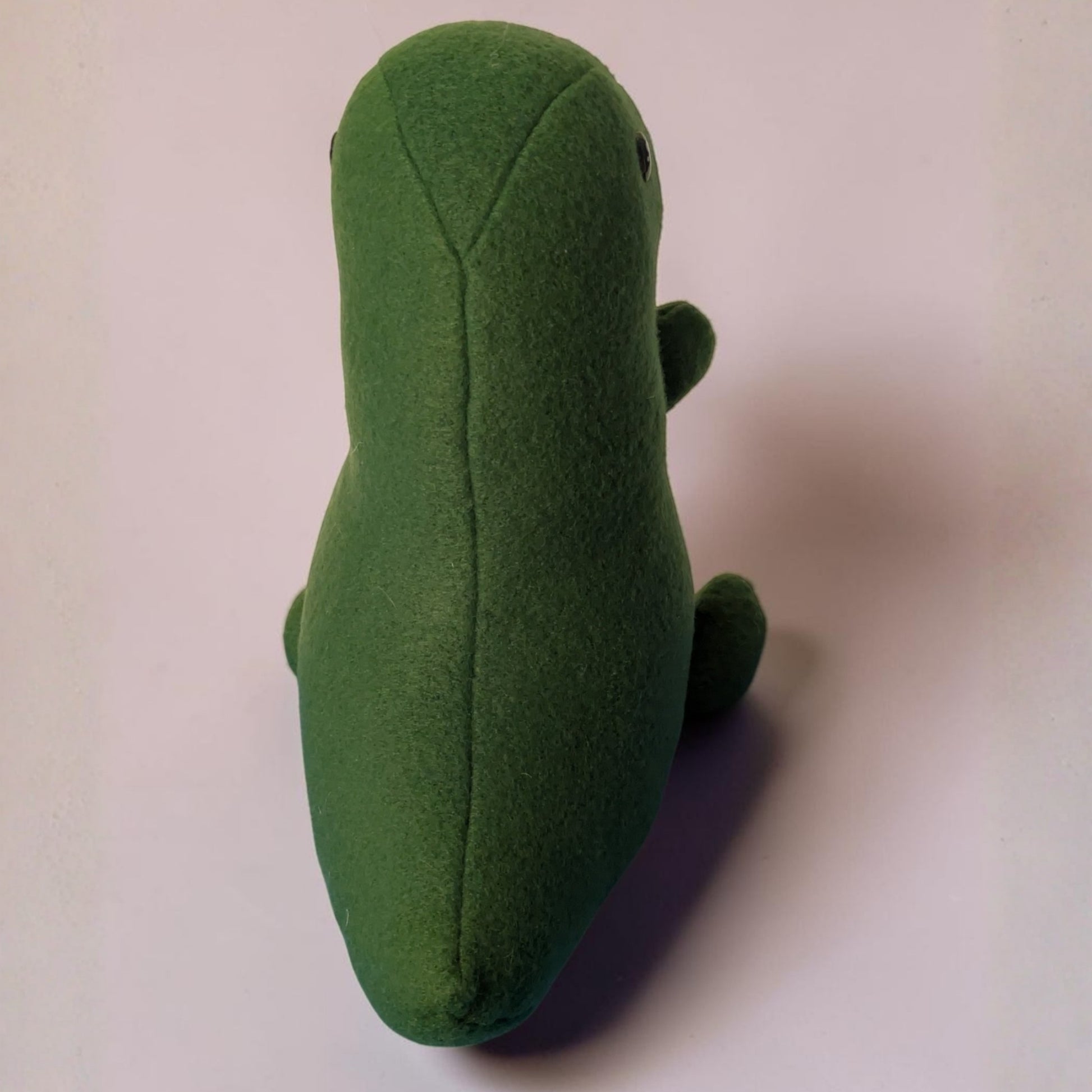 A green plush toy shaped like a stylized dinosaur, photographed from the back on a plain light background.