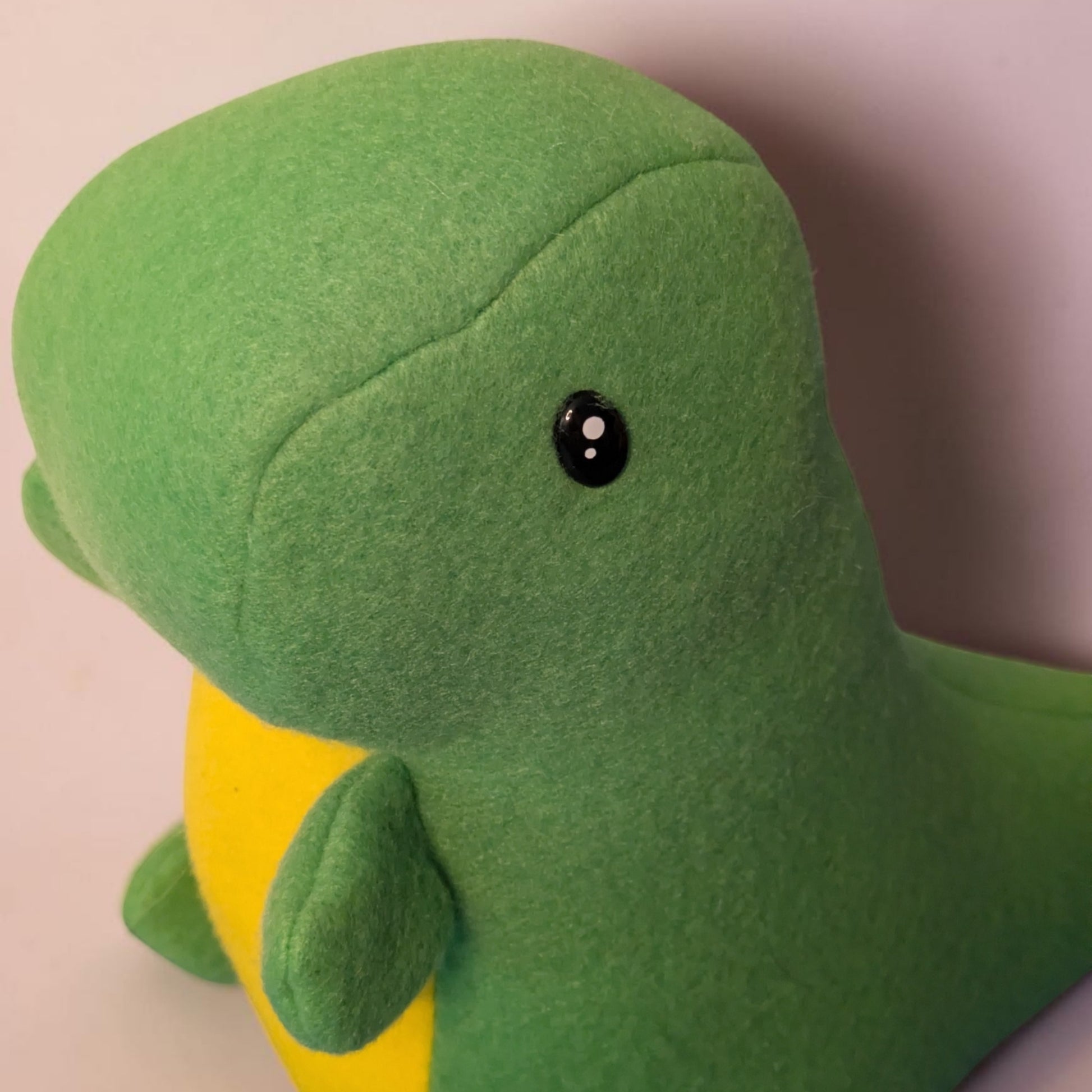 A close-up of a green plush dinosaur toy with a yellow belly and a black safety eye, photographed against a plain light background.