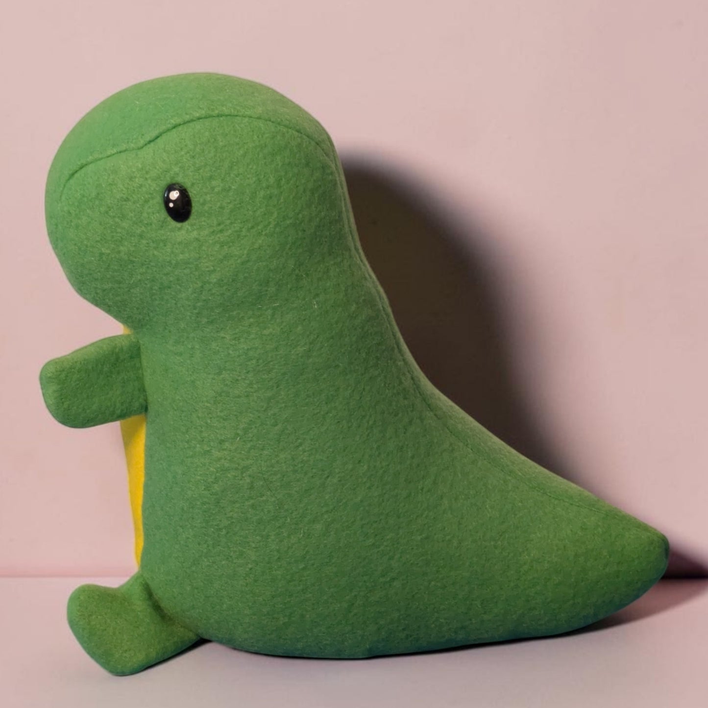 A green fleece dinosaur plush toy with a small yellow belly patch and black safety eyes, sitting against a light pink background.