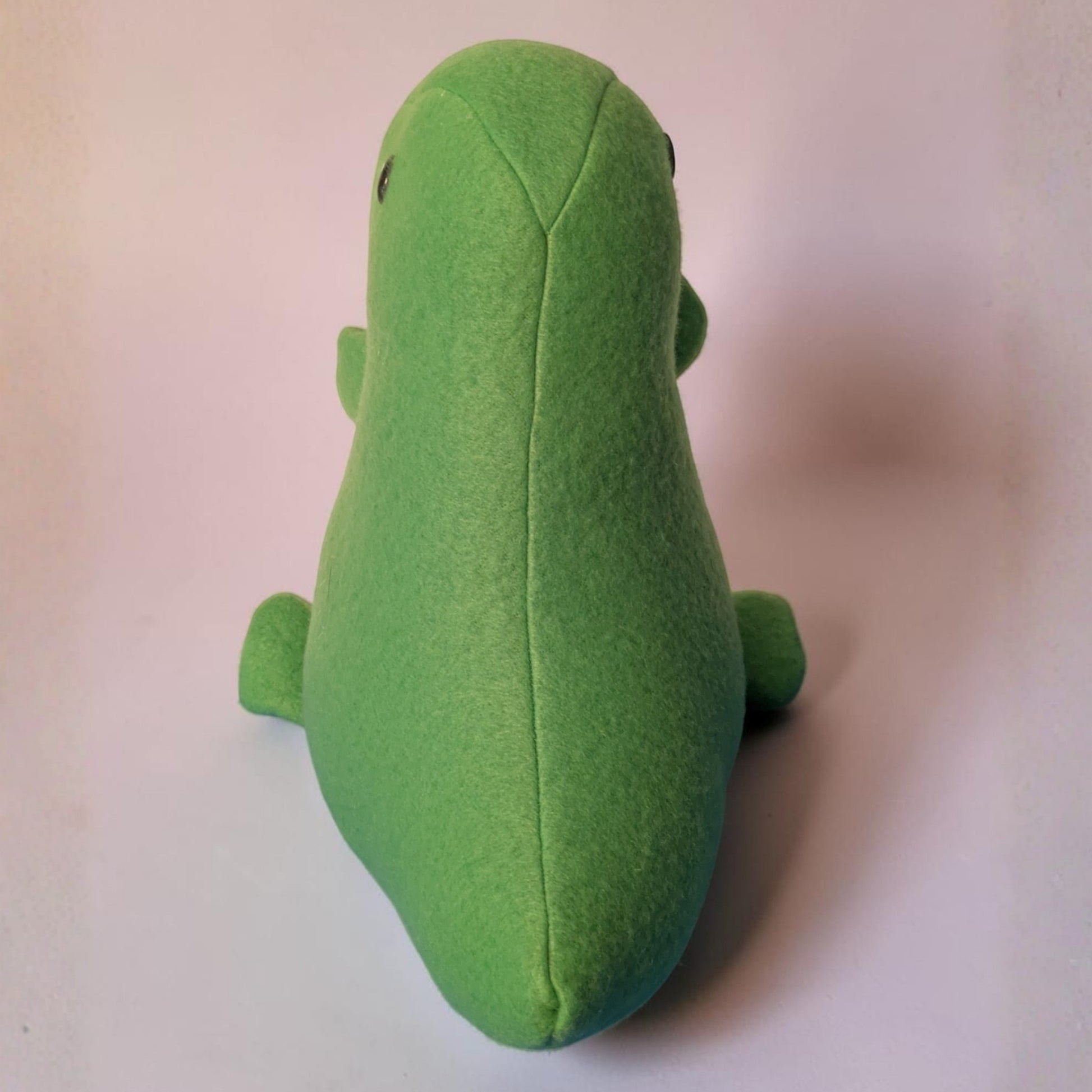 A green plush toy shaped like a stylized dinosaur, photographed from the back on a plain light background.