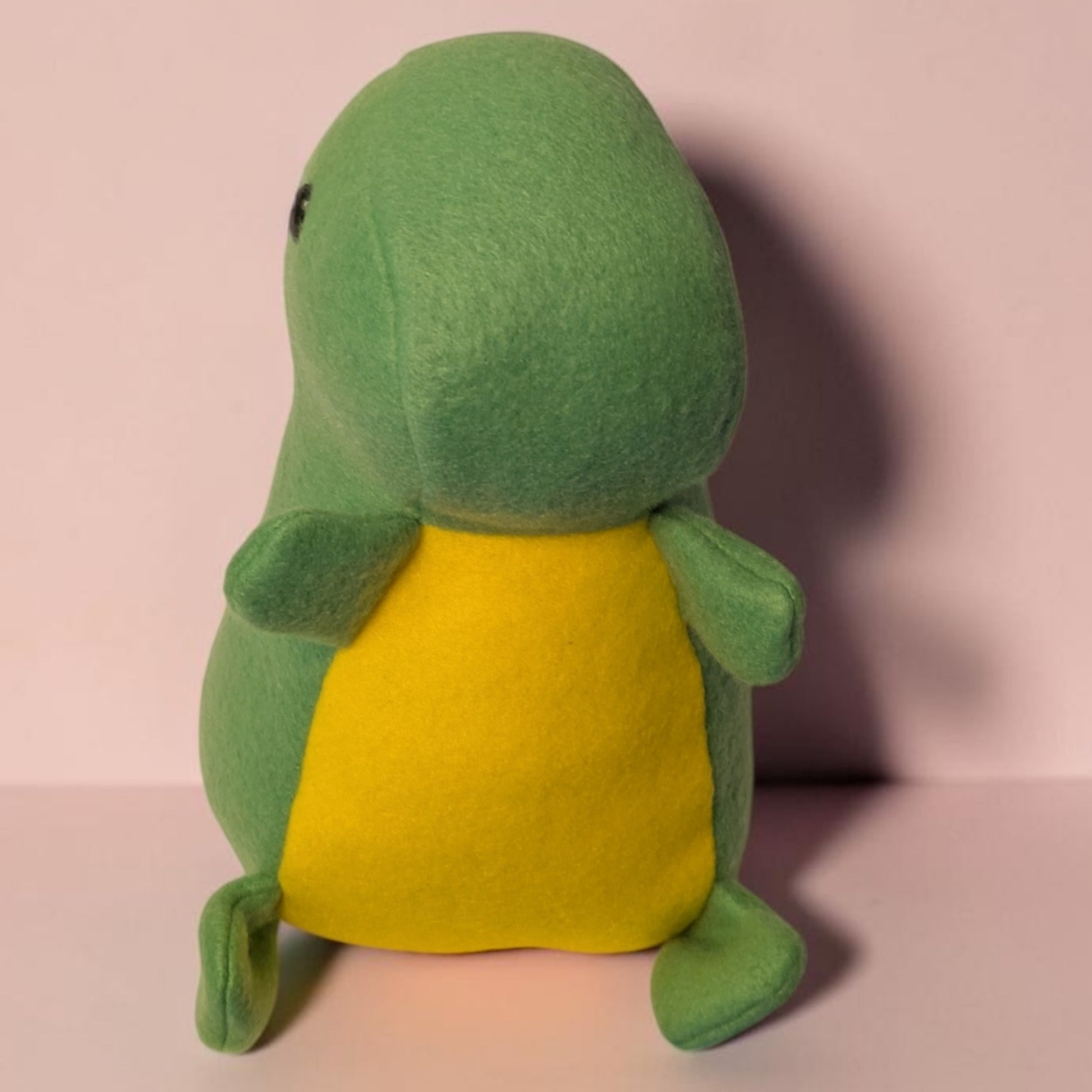 A green plush dinosaur toy with a yellow belly, photographed against a soft pink background.