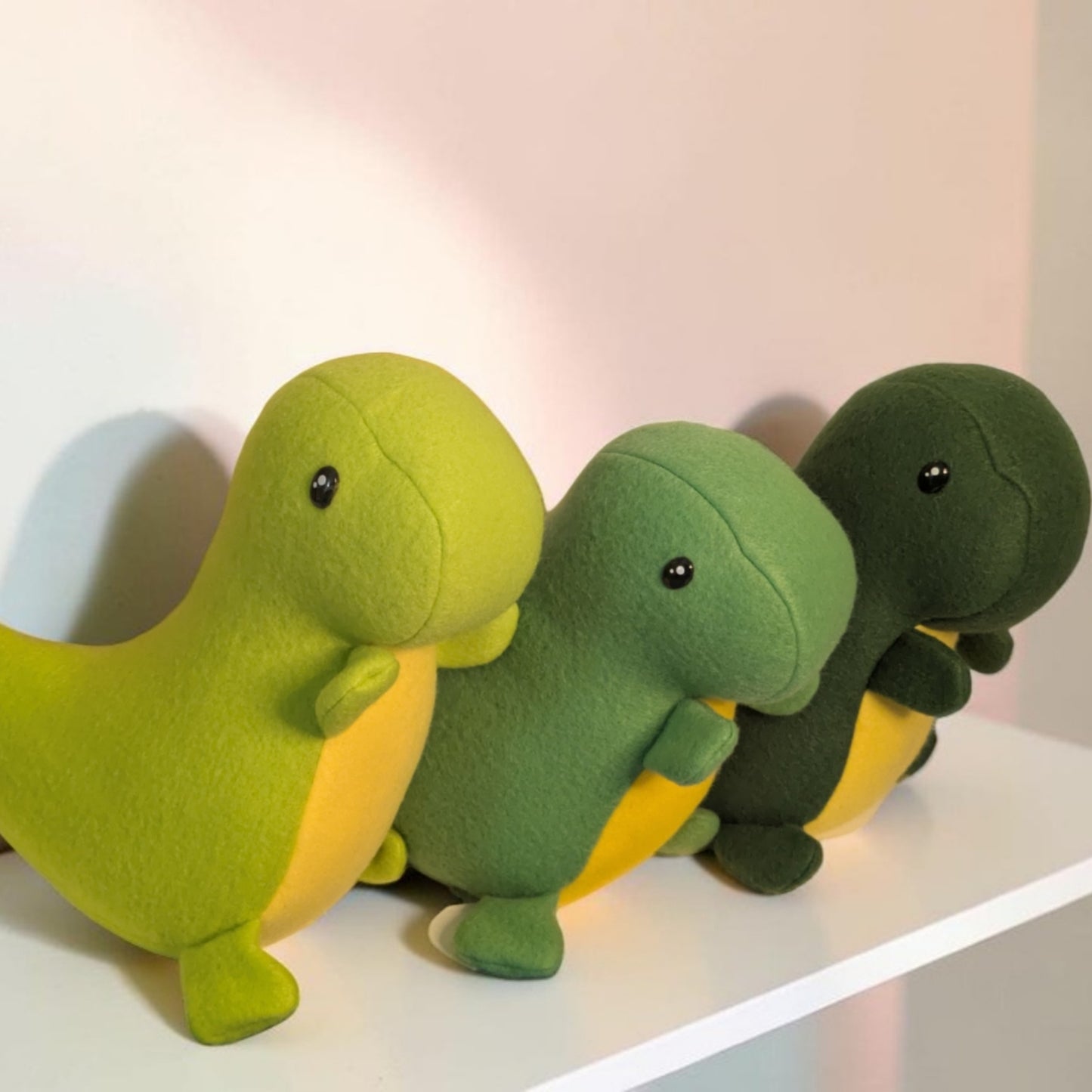 Three plush dinosaur toys lined up on a white shelf against a light wall. The dinosaurs are lime green, medium green, and dark green, each with yellow underbellies and small black safety eyes.