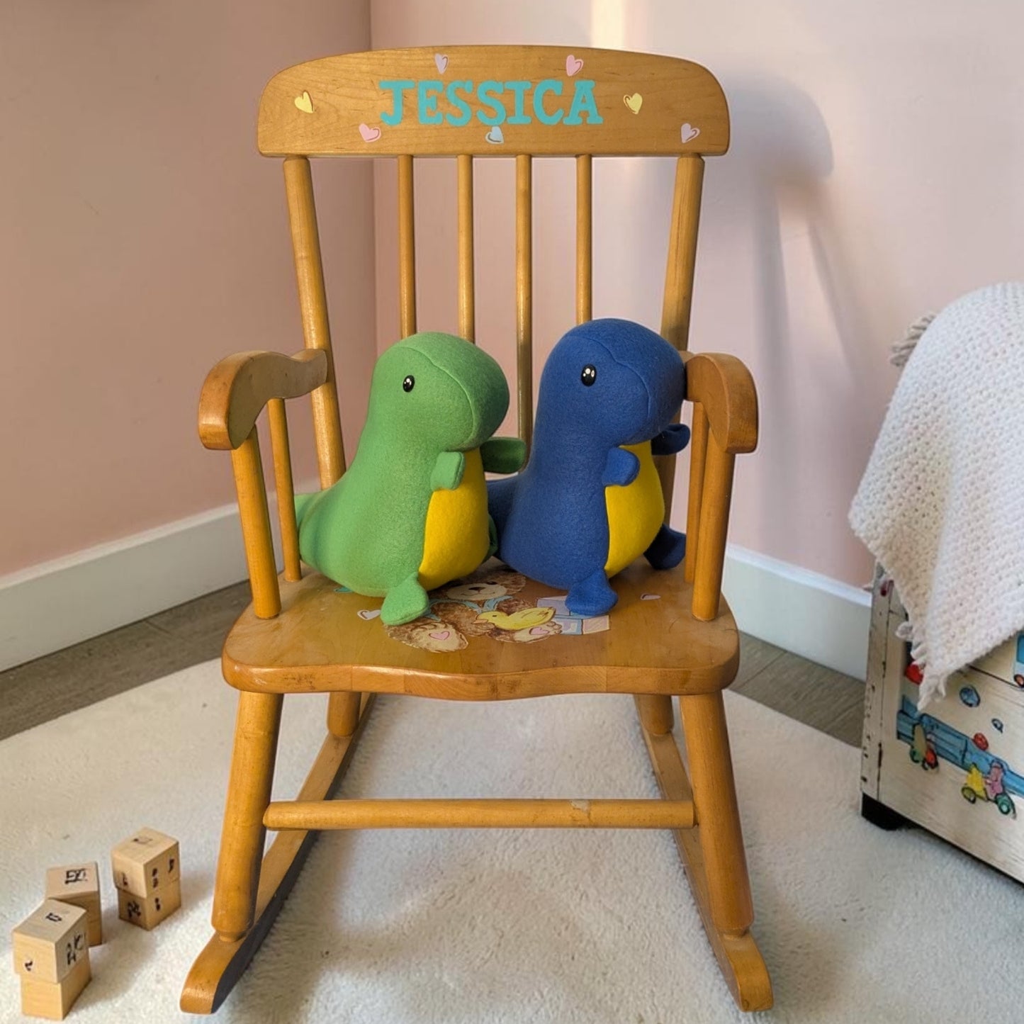 A wooden rocking chair in a nursery setting, featuring the name 'JESSICA' personalized on the backrest. Two plush dinosaur toys, one green and one blue, are sitting on the chair.