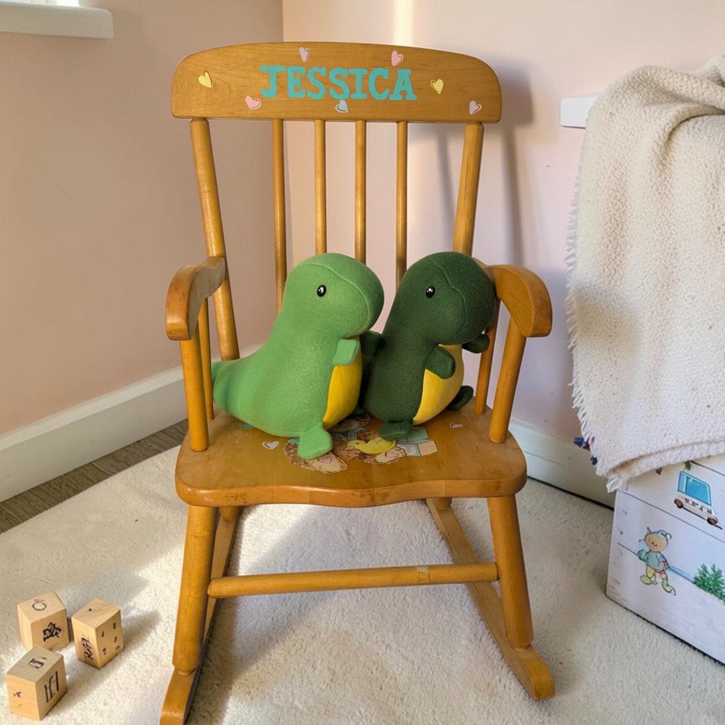A wooden rocking chair with the name 'JESSICA' painted on the backrest, styled in a nursery with two plush green dinosaur toys on the seat.