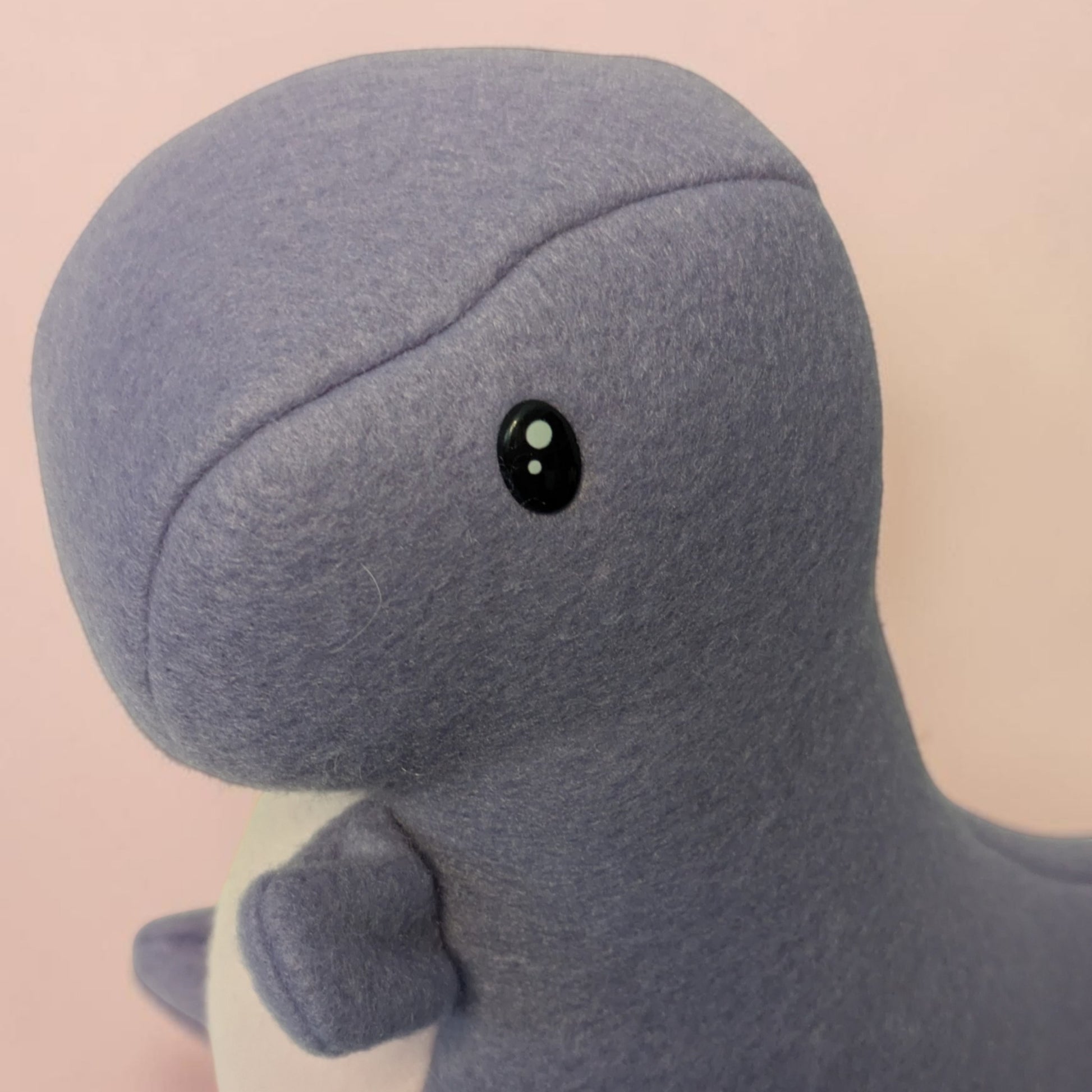 A close-up of a plush dinosaur toy made of soft textured lavender fleece, featuring a small black safety eye with white highlights, set against a soft pink background.