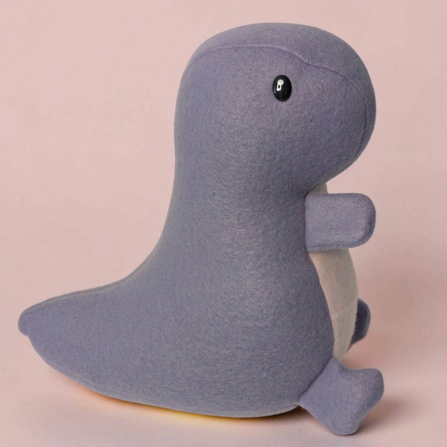 A plush Dinosaur toy made of soft lavender fleece fabric with a small black safety eye and a pale pink belly, standing against a solid pale pink background.