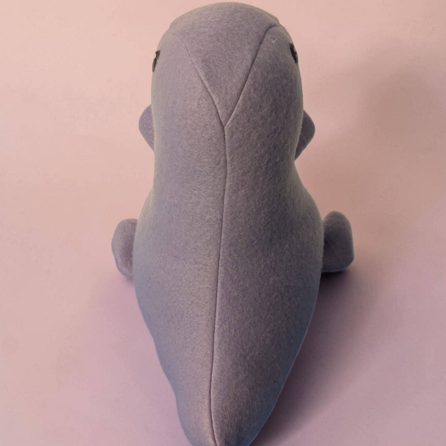 A lavender purple plush dinosaur toy photographed from the back against a soft pink background.