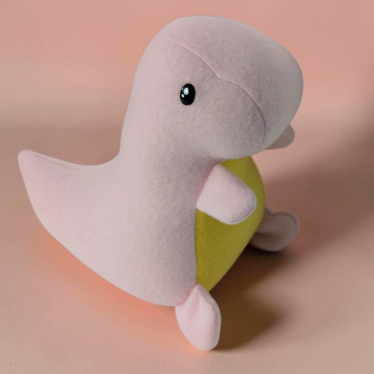 A plush toy shaped like a dinosaur, with a pale pink body, yellow belly, and small black eyes, photographed on a soft peach-pink background. 