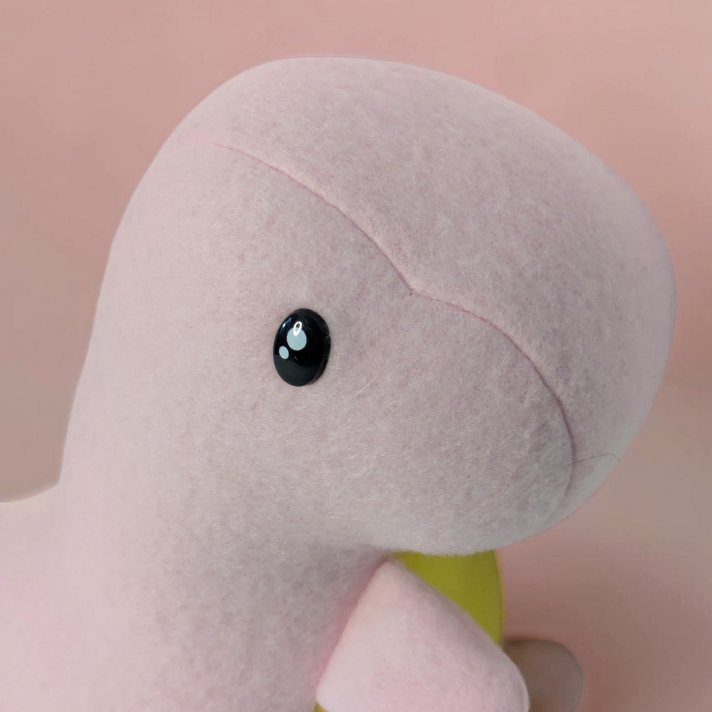 A close-up of a soft pink plush dinosaur toy with a black safety eye and a small yellow belly, set against a solid pale pink background.
